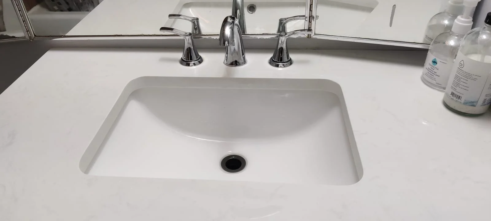 Bathroom white sink with three chrome faucets, mirror above, and soap dispensers on the right side of the countertop.