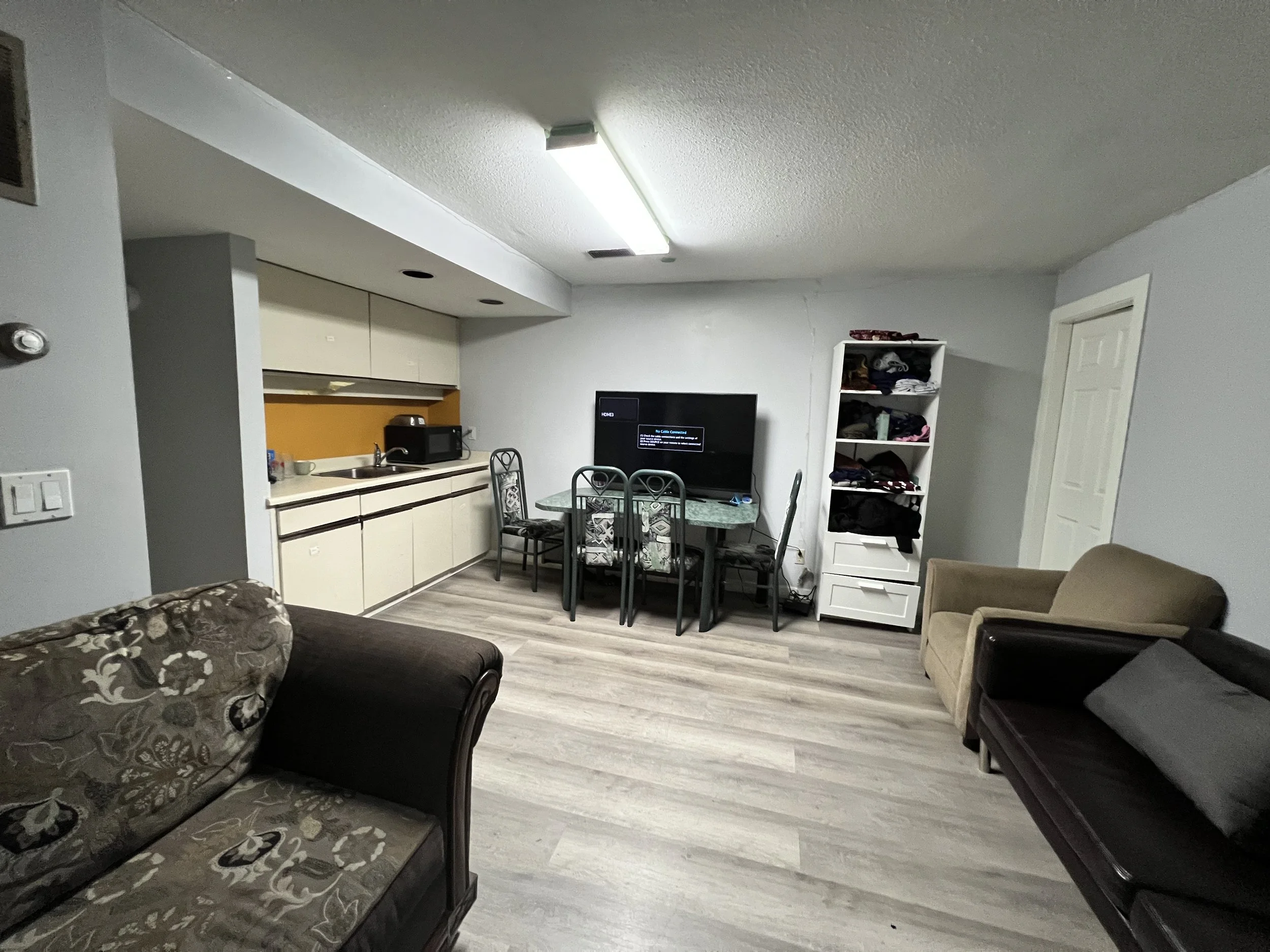 Living room with a TV on a stand, a glass dining table with chairs, a small kitchen area to the left, and sofas on the right side with a bookshelf filled with clothes and items against the wall.