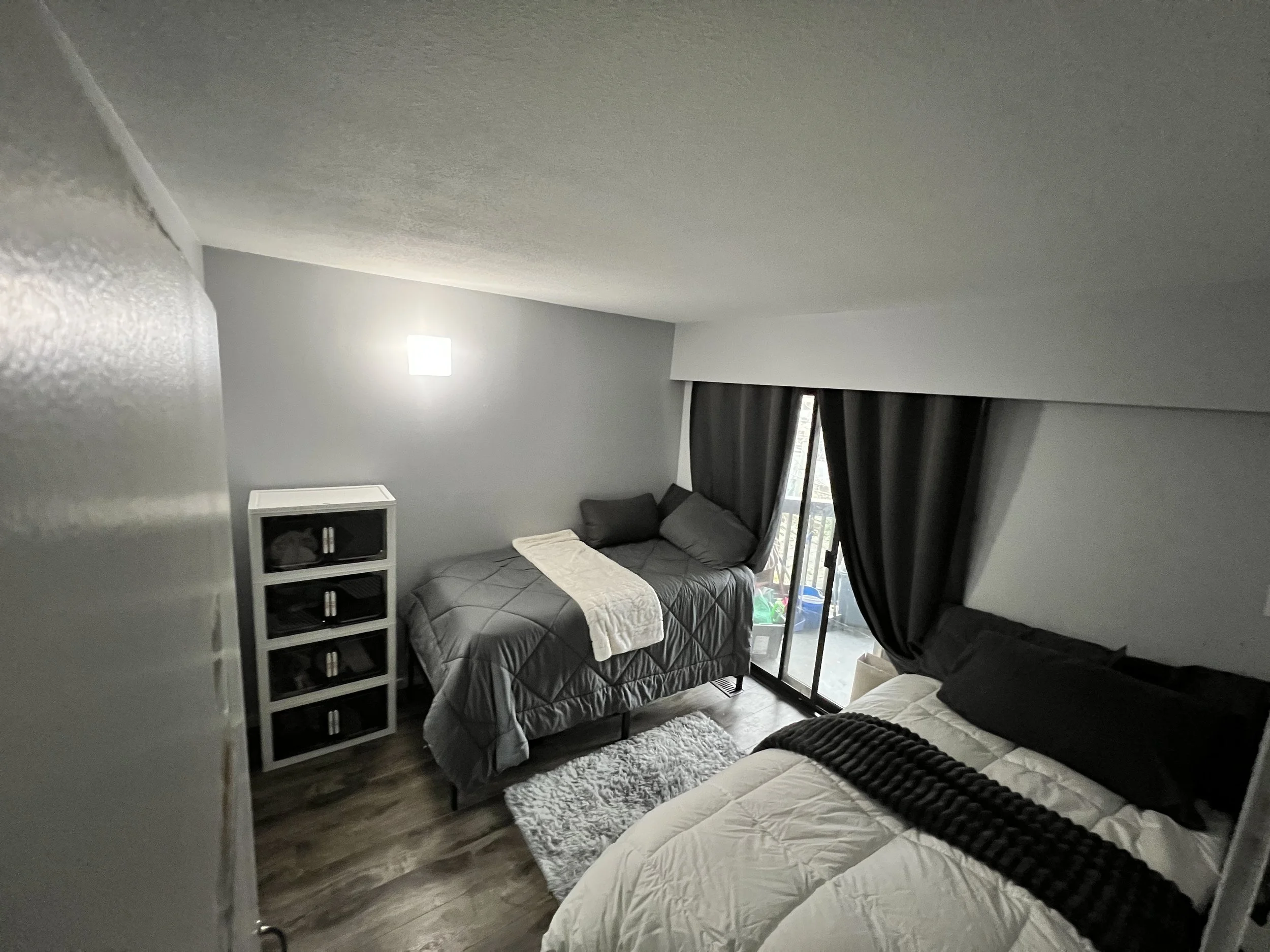A bedroom with two beds, one with dark gray bedding and one with white bedding, a small white shelving unit, a window with dark curtains, and a small rug on wooden flooring.