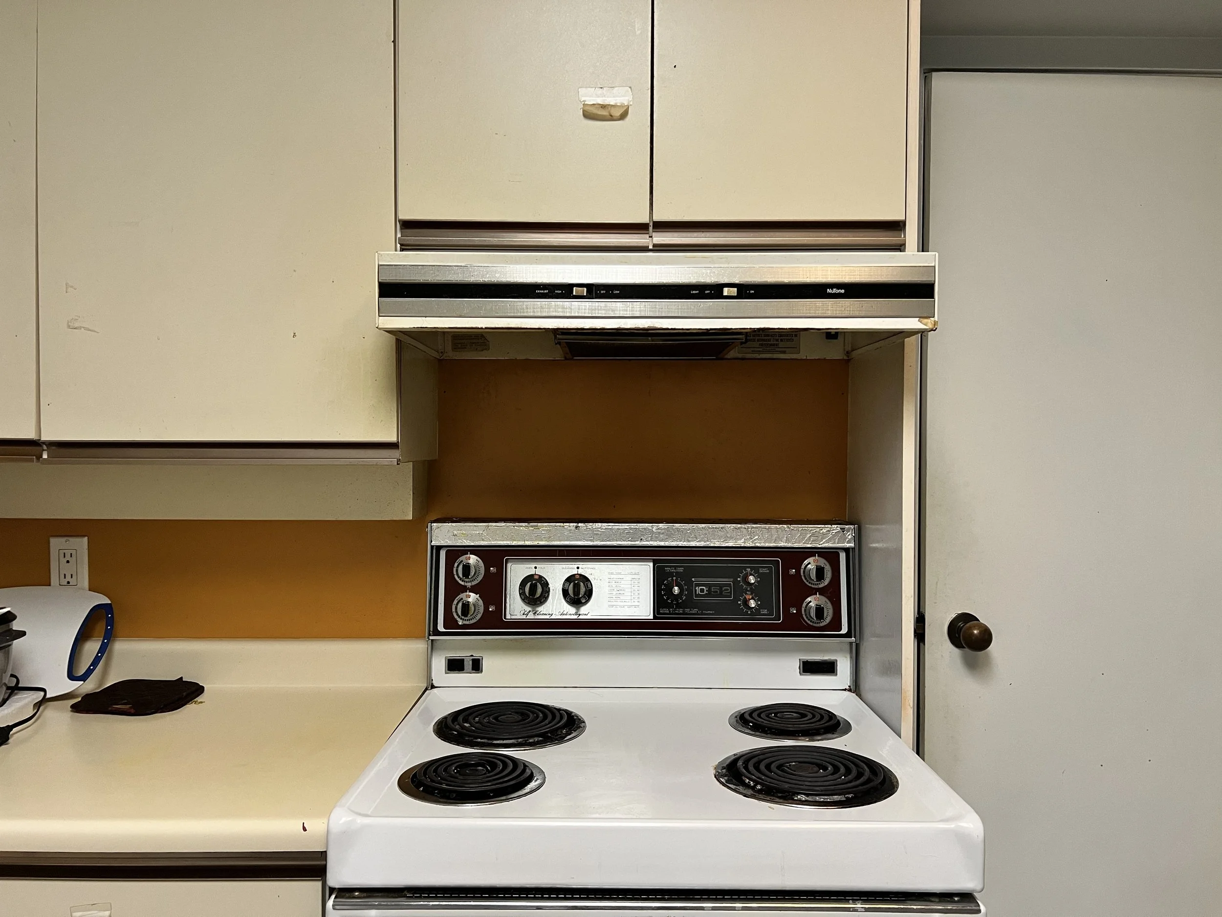 An old kitchen stove with four coil burners, a control panel with dials, and a vent hood above. To the left, there is a white countertop with a small black item, a jar, and an electric outlet. To the right, there is a beige door with a round knob.