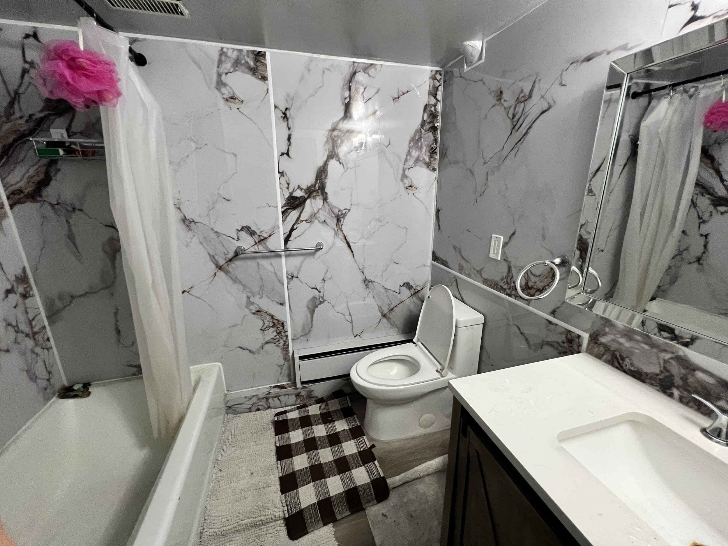 Bathroom with marble walls, a shower with a curtain, a toilet, a large mirror, a sink with a vanity, a towel ring, and a small rug.