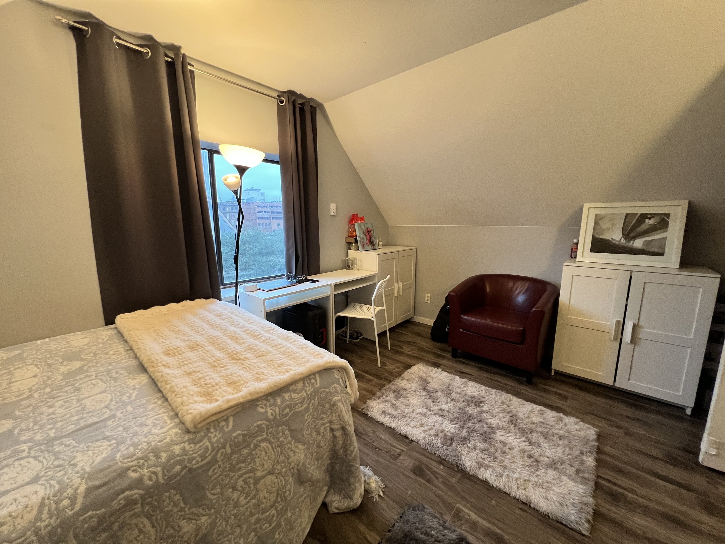 A cozy bedroom with a bed covered in a patterned beige comforter and a cream-colored blanket on top. There is a window with dark curtains, a tall floor lamp, a small white desk with a chair, a white storage cabinet, a red armchair, a shaggy rug on wo