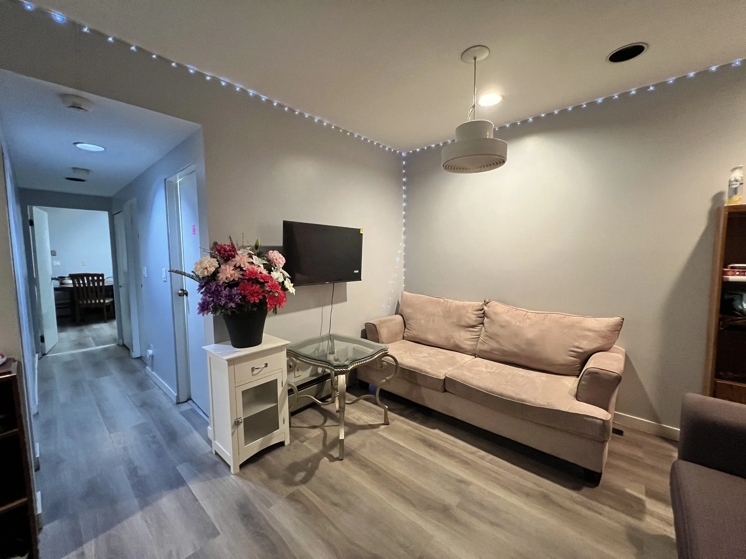 Living room with a beige sofa, glass coffee table, wall-mounted TV, a small white side table with a large flower vase, string lights on the ceiling corner, and a wooden shelf on the right. A hallway leads to other rooms in the background.