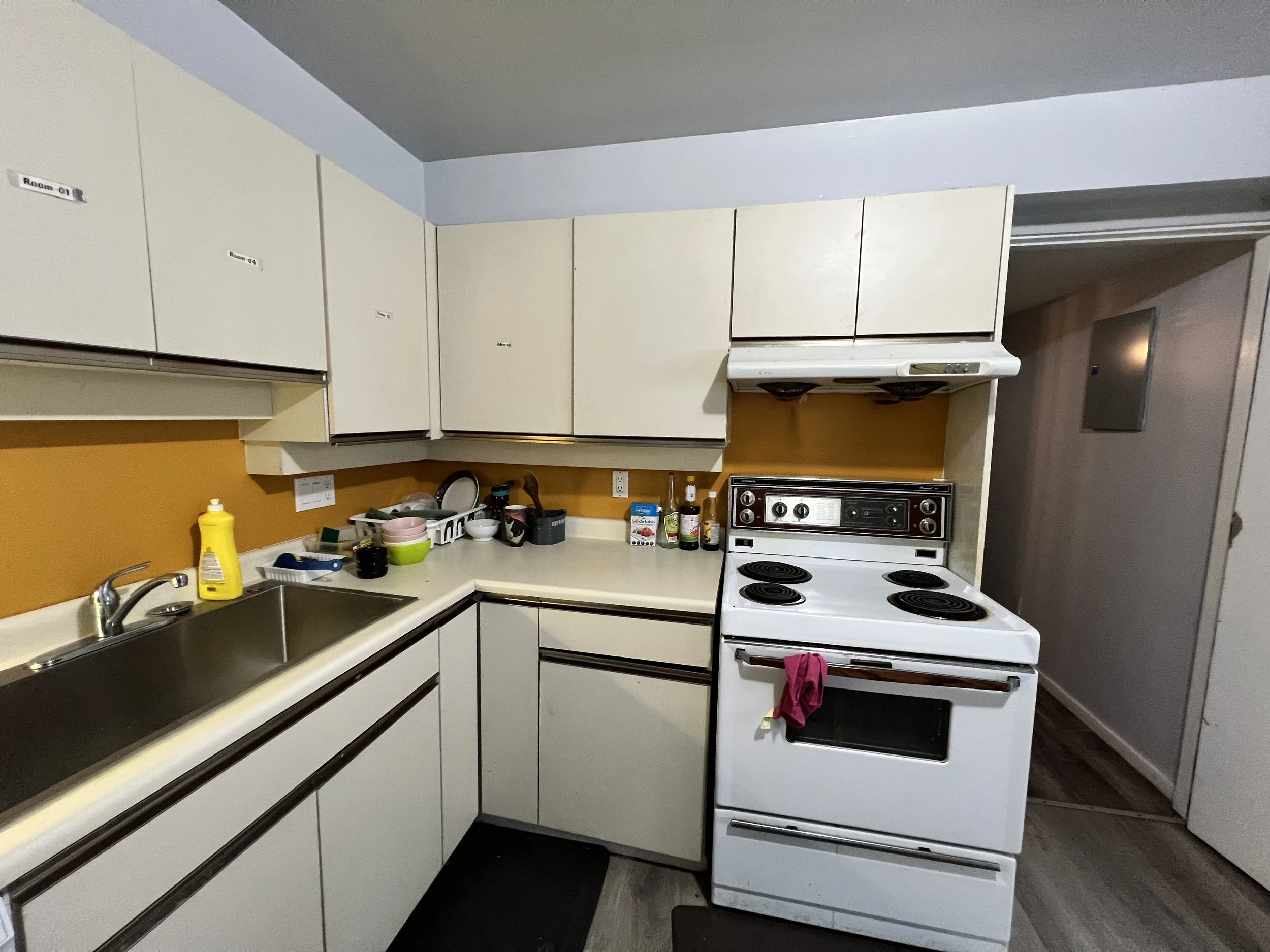 Kitchen with white cabinets, yellow wall, black stovetop and oven, and various bottles, dishes, and cleaning supplies on the counters.