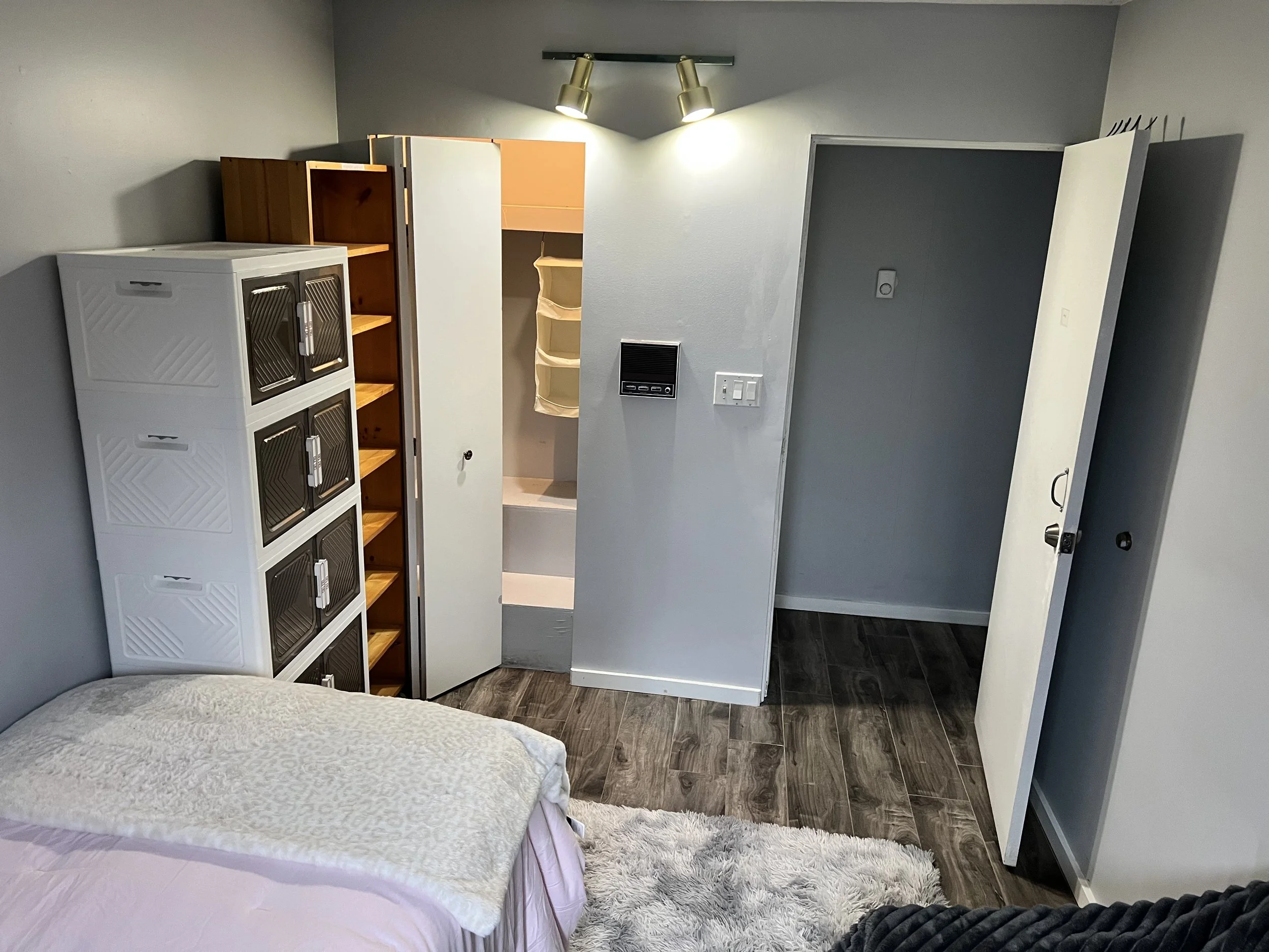 A bedroom with a bed, a white storage unit with black doors, an open closet with hanging organizers, a light switch, and wood flooring.
