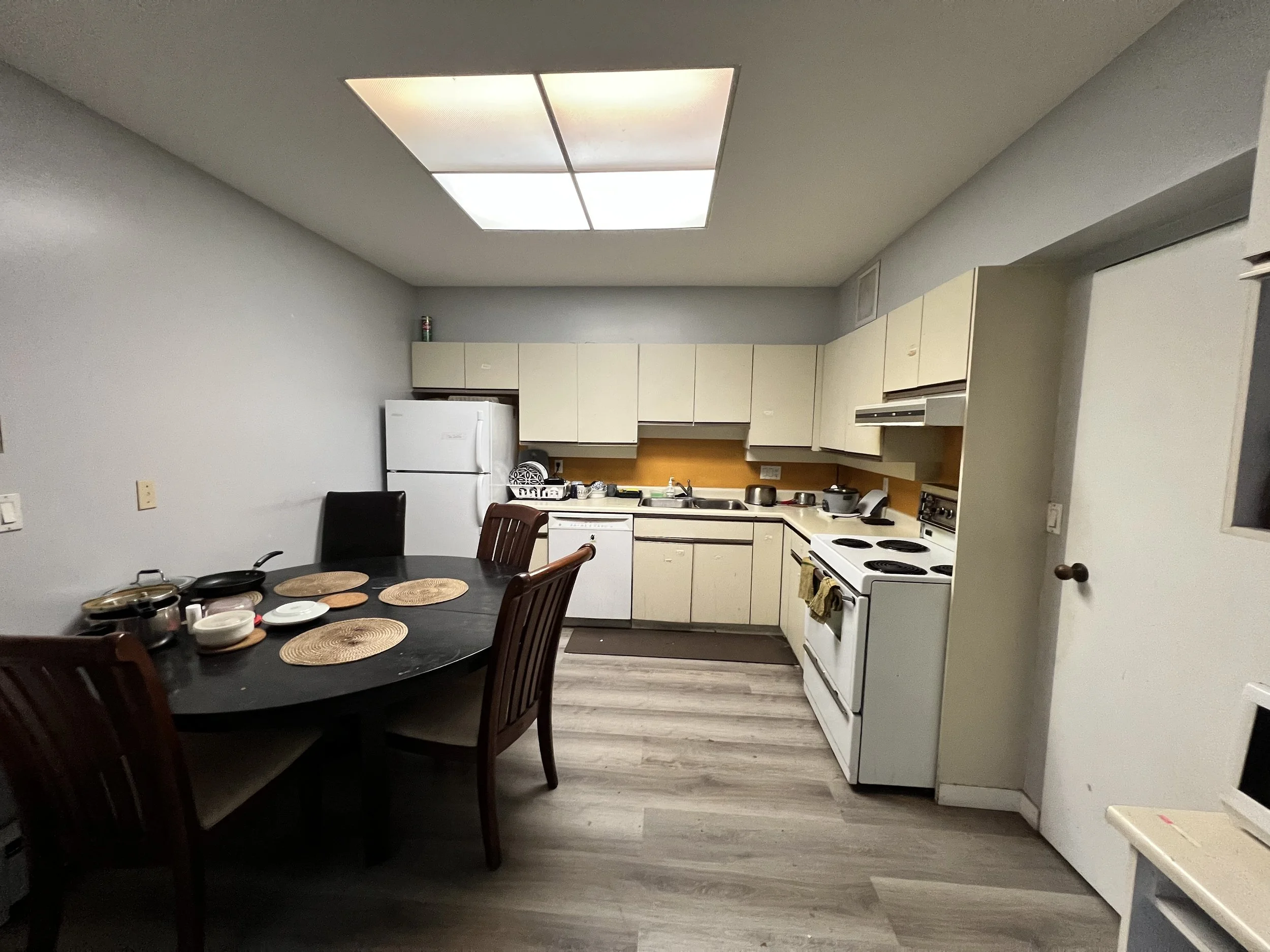 Kitchen with white cabinets, white refrigerator, white stove, and a black dining table with wooden chairs, some dishes and kitchen utensils on the table, and a ceiling fluorescent light