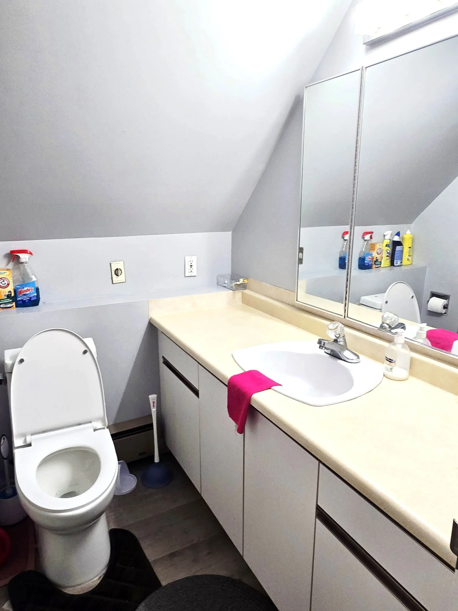 A small bathroom with a toilet and a white vanity with a sink, pink cloth on the counter, and cleaning supplies above the toilet, slanted ceiling, mirror cabinets above the sink, and wall outlets.