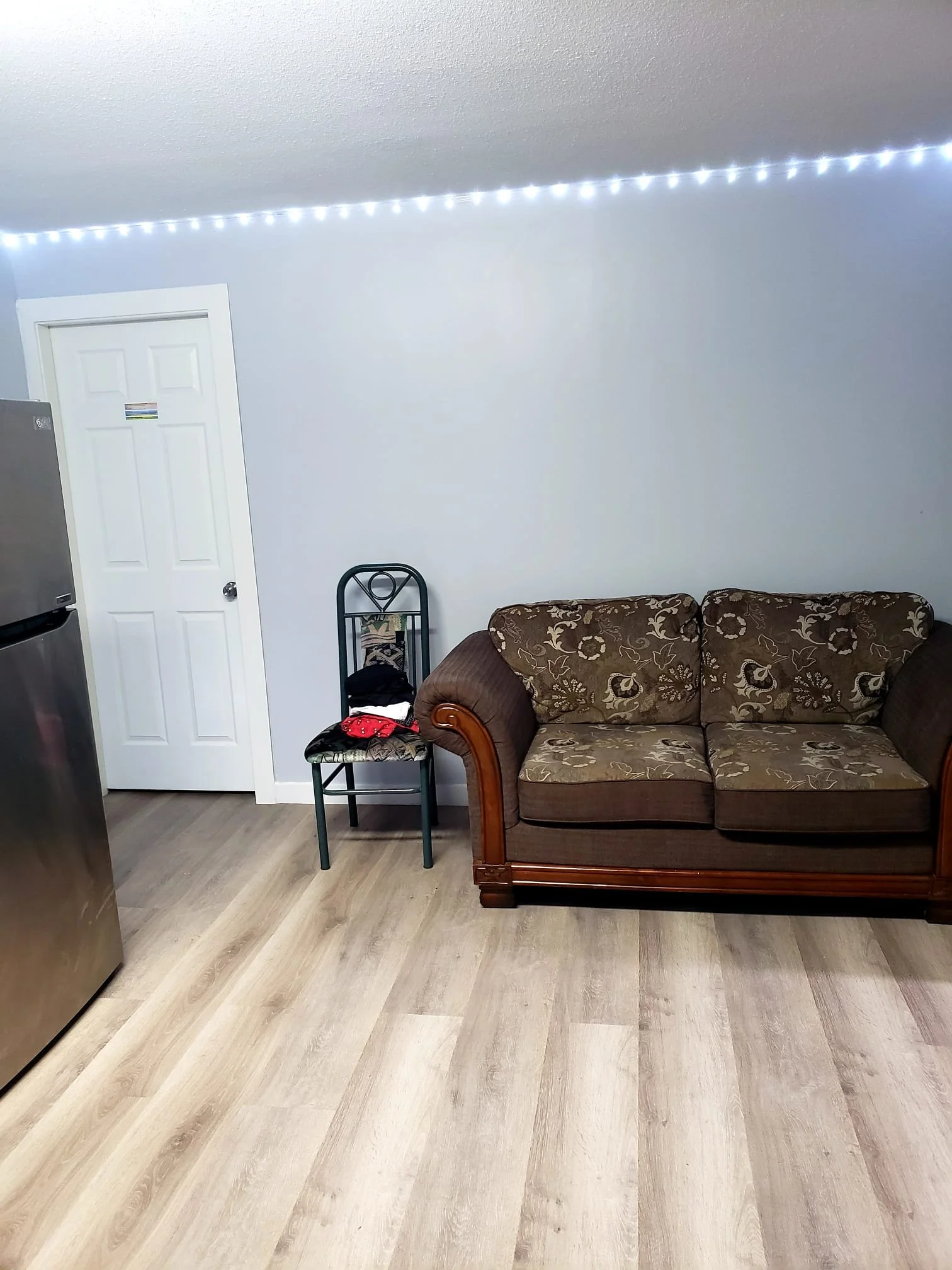 Living room with a brown floral-patterned couch, a black chair, a white door, a refrigerator, and a string of white LED lights on the ceiling.