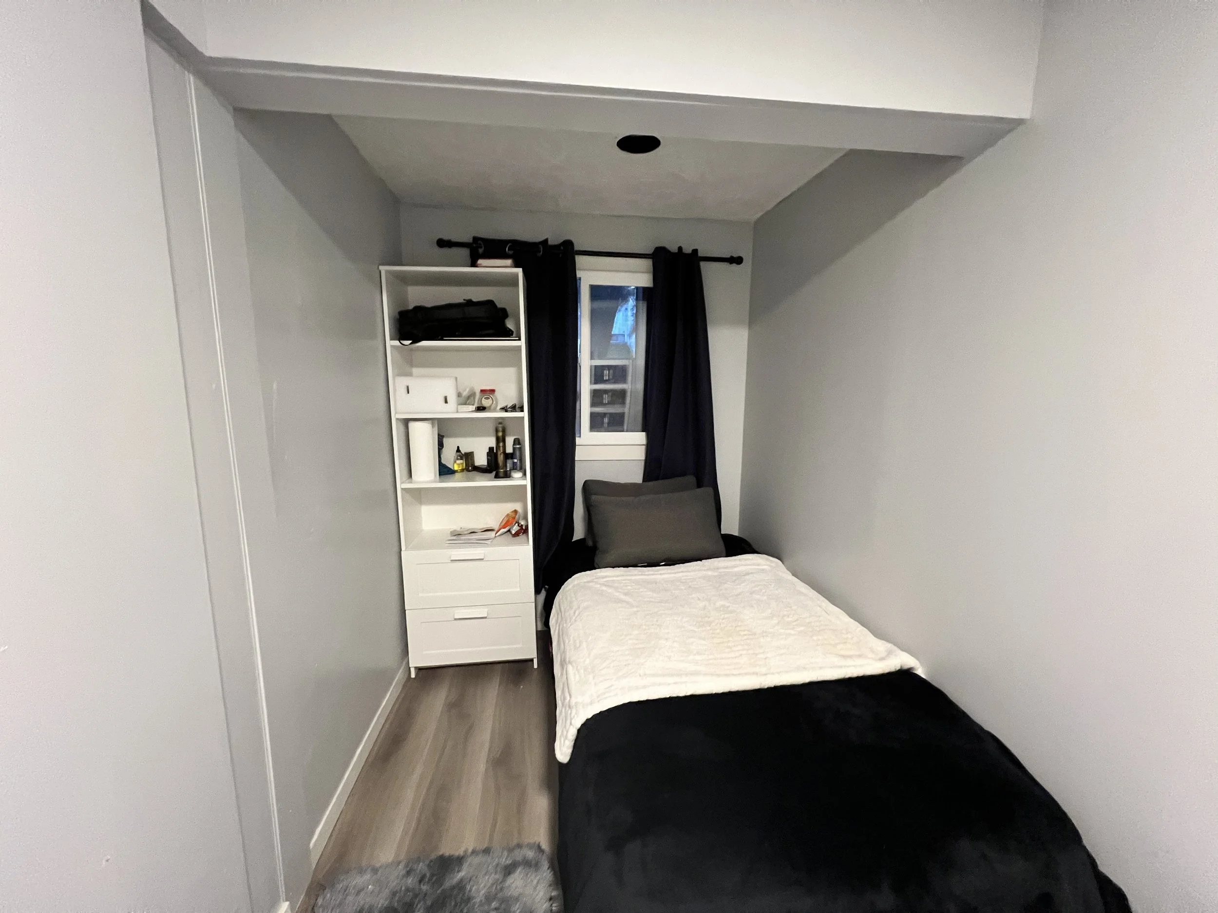 Small bedroom with a single bed, white quilt, and dark bedding. Small window with black curtains, white shelving unit with various items, and gray walls.
