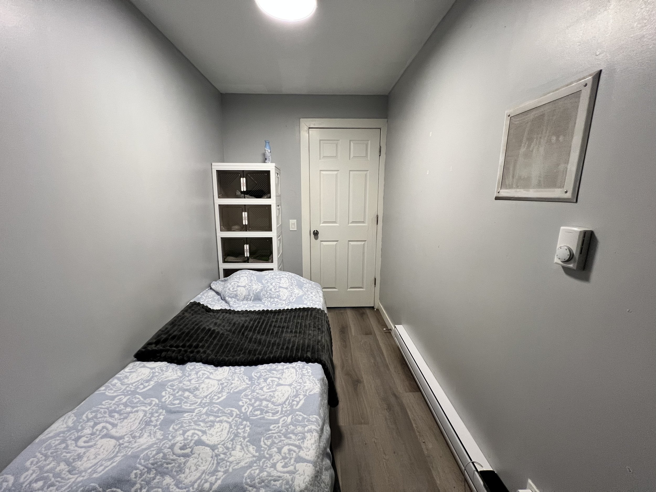 Small bedroom with a single bed covered in white and gray bedding, black throw blanket, gray walls, a white cabinet with mesh doors, a door at the end of the room, a wall vent, thermostat, and a baseboard heater.