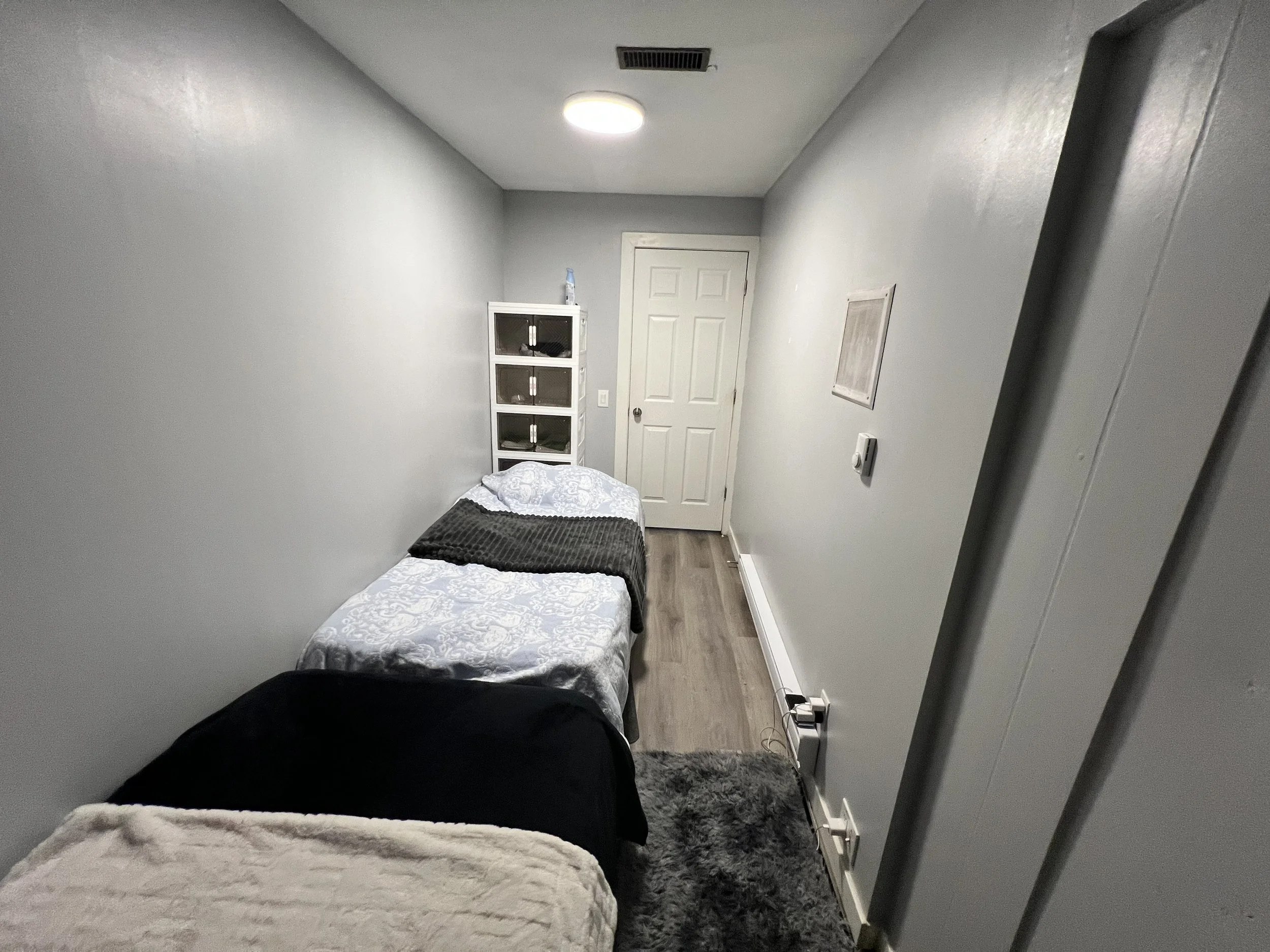 Small room with a bed on the left side, a white shelf with glass doors, a door at the far end, and a gray carpet on the floor.