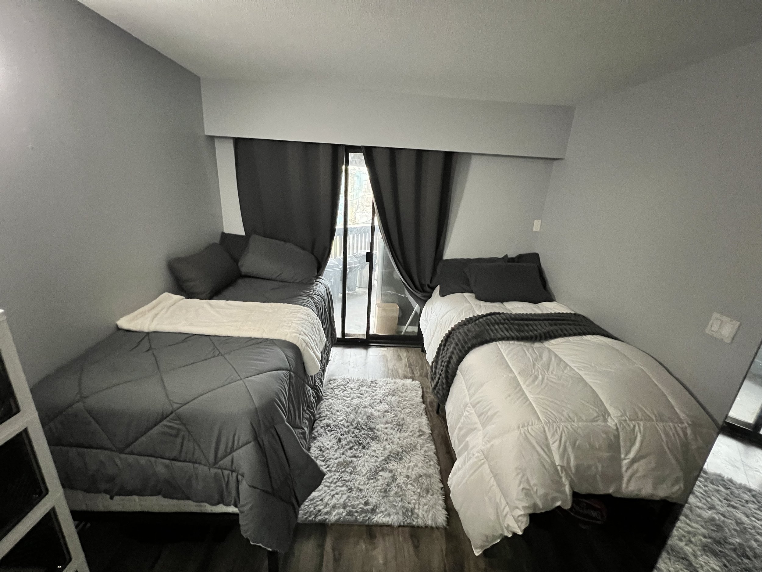 A small bedroom with two beds, one on each side of a sliding glass door leading to a balcony, with dark curtains, gray and white bedding, and a textured white rug.