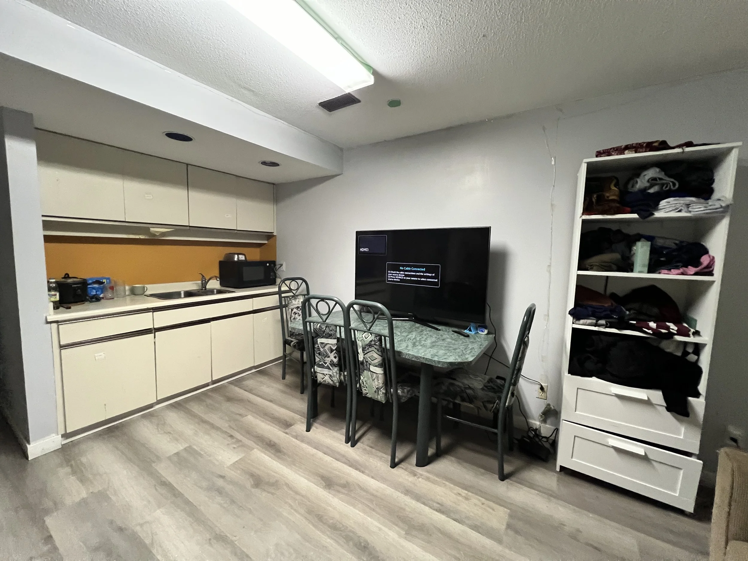 Living room with kitchenette, table with chairs, and stacked shelving with clothes, TV displaying no signal message.