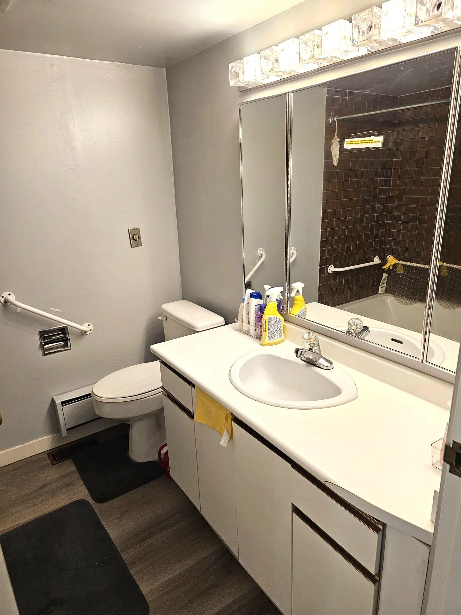 A small bathroom with a white sink vanity, towels, cleaning supplies, a large mirror, and a shower area with brown tiles and a grab bar.