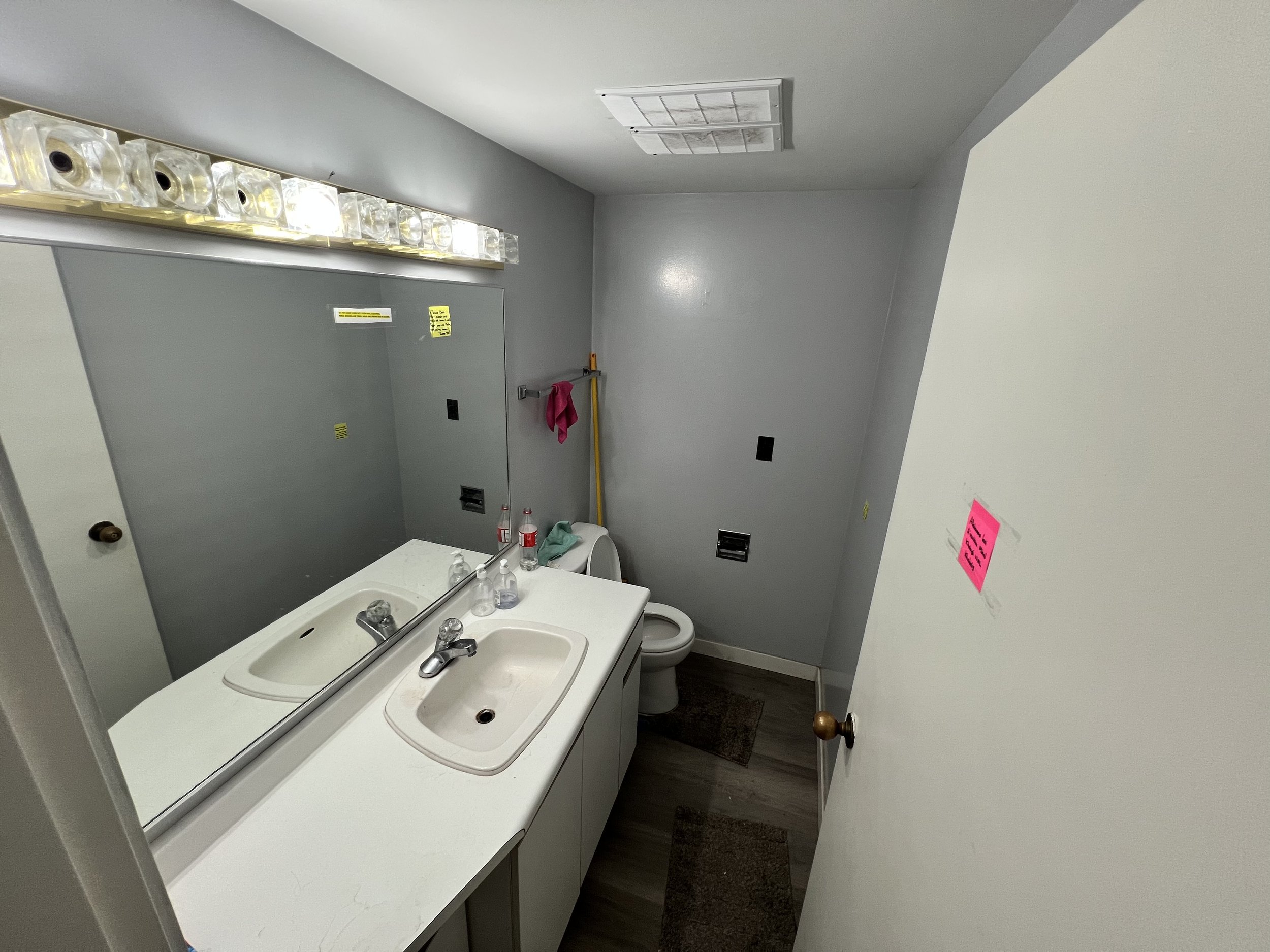 Small bathroom with a double sink vanity, mirror, fluorescent light, toilet, gray walls, and brown floor mats.