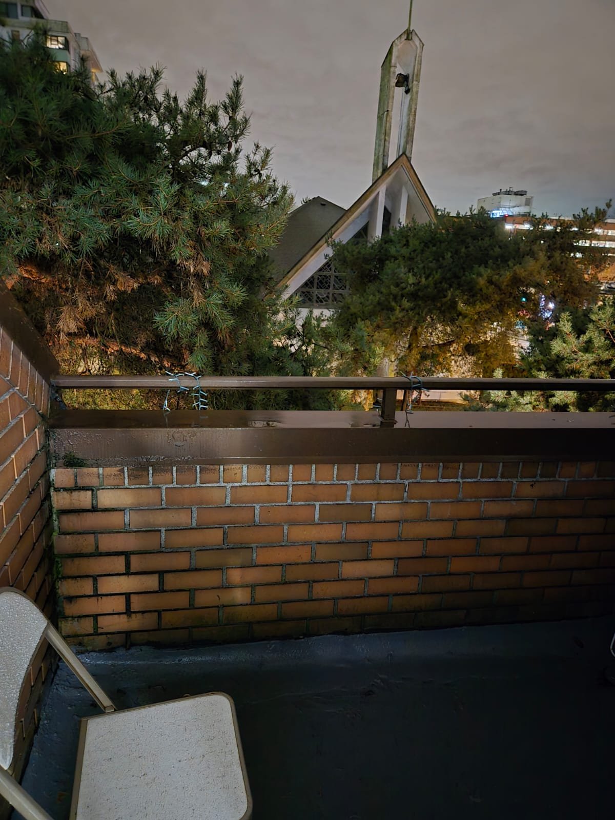 View of a small balcony with a brick wall, a metal railing, and two outdoor chairs. Behind the balcony, there are trees and a church-like building with a tall steeple. The sky is overcast.