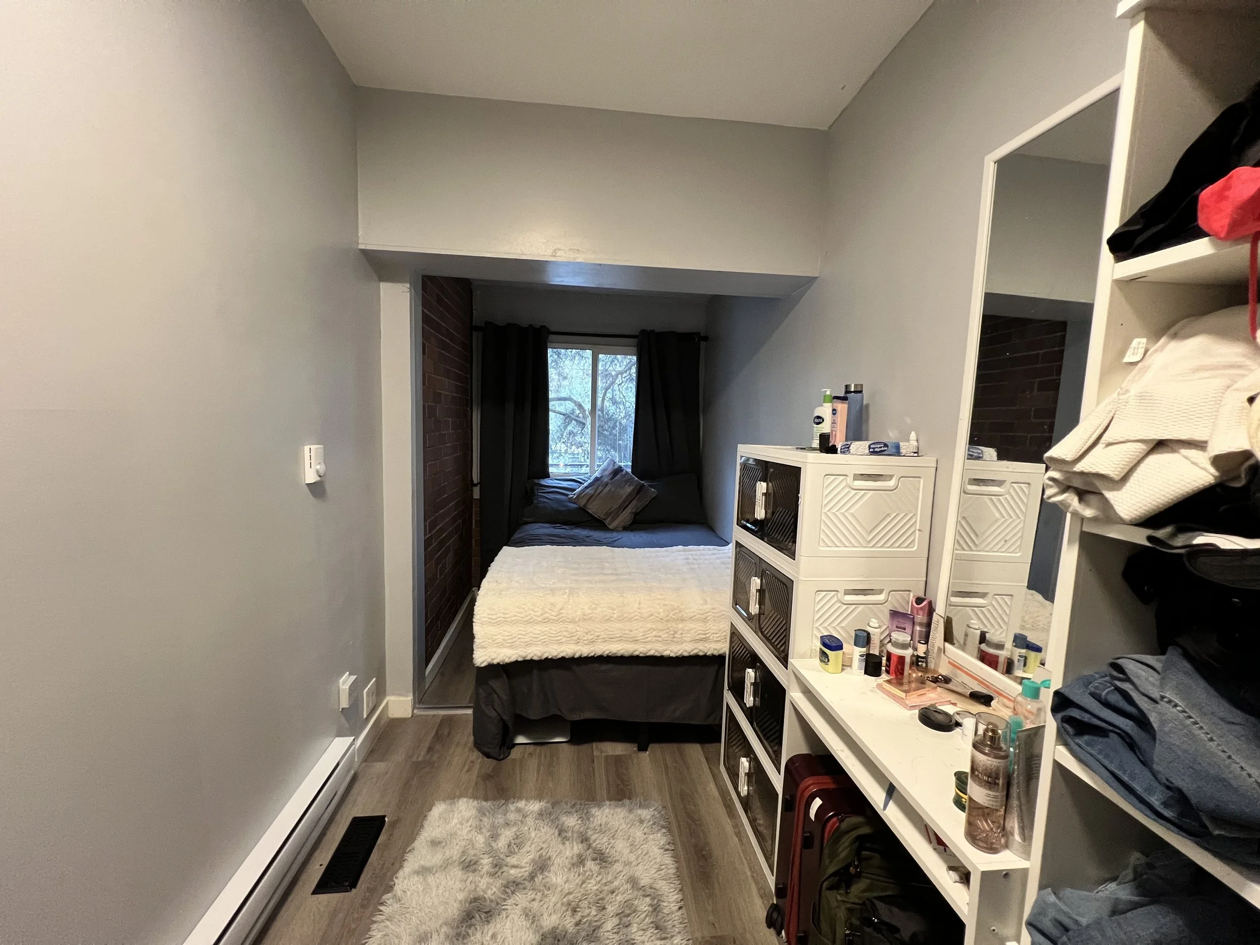 Small bedroom with bed near window, white dresser with toiletries, mirror, and shelves with clothes, hardwood floors, gray walls, black curtains, and a beige rug.