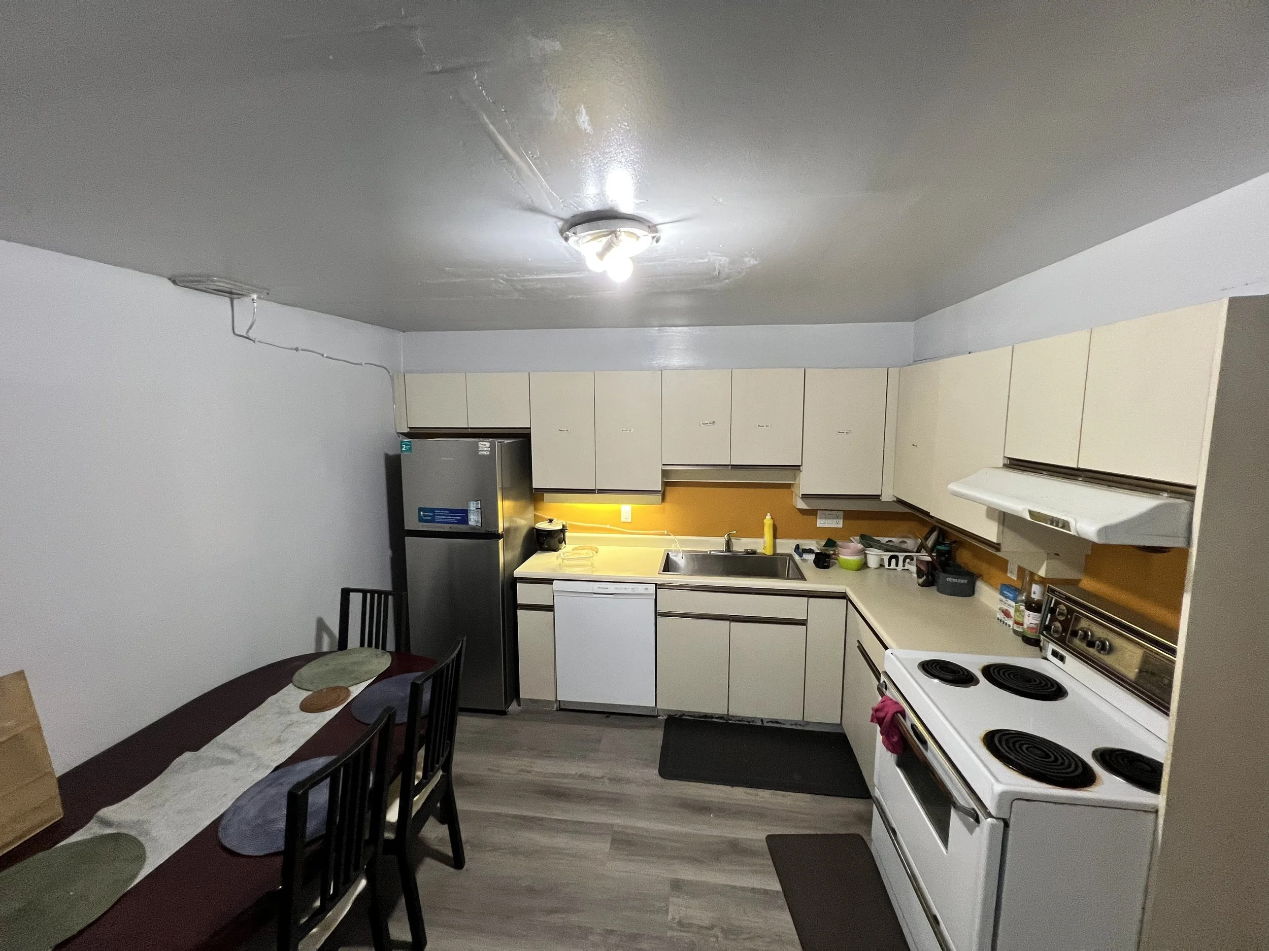 Small kitchen with white cabinets, a gray refrigerator, a white electric stove, and a dining table with black chairs. The table has placemats and a paper bag. The kitchen has gray flooring and a yellow backsplash.