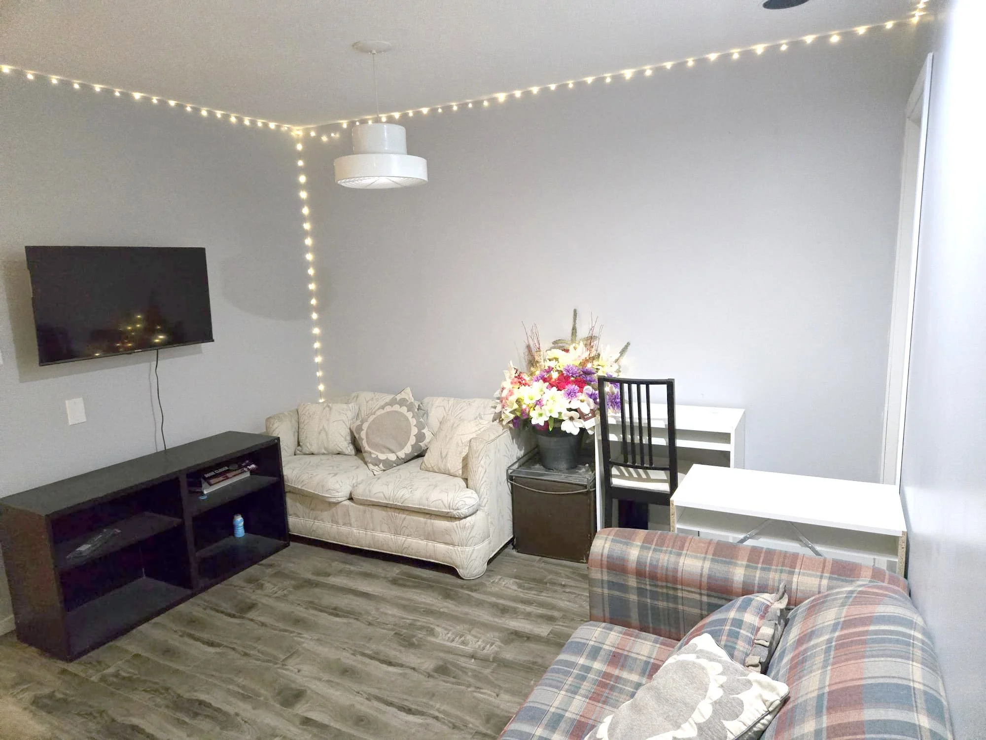 A living room with a wall-mounted flat-screen TV, a beige sofa with pillows, a plaid armchair, a black flower pot with colorful flowers, a white desk, and string lights on the ceiling and corner.