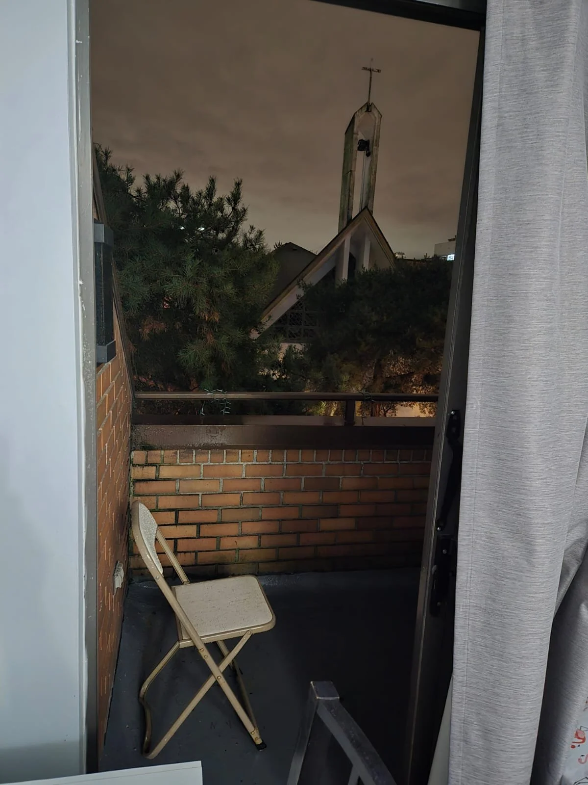 Nighttime view of a church steeple through a window, with a small balcony and a folding chair inside the room.