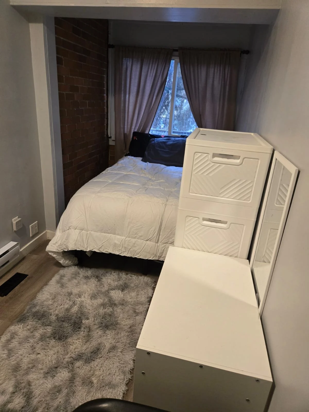 A small bedroom with a bed, window with closed curtains, a white storage unit, a mirror, and a grey fluffy rug on the wooden floor.