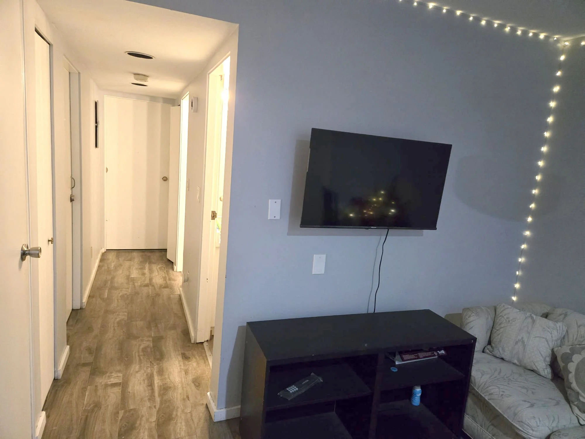 Furnished living room with wall-mounted TV, string lights, and a small sofa with pillows. Hallway with closed doors leading to other rooms.