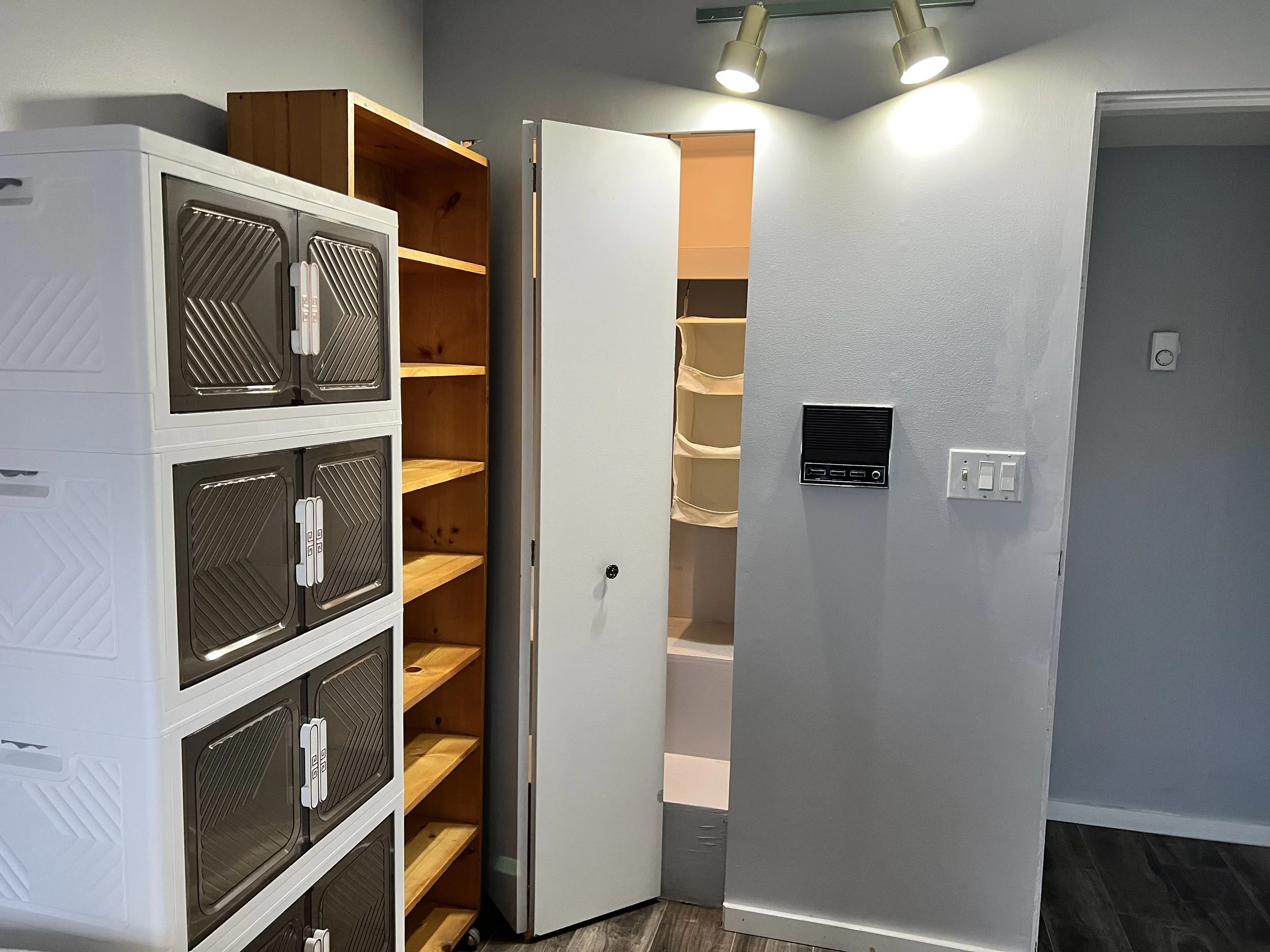 A laundry room with six white plastic storage units stacked vertically, a wooden bookshelf with five empty shelves, an open door revealing a storage closet, a wall-mounted small black speaker, and a light switch panel. The ceiling has two spotlights,