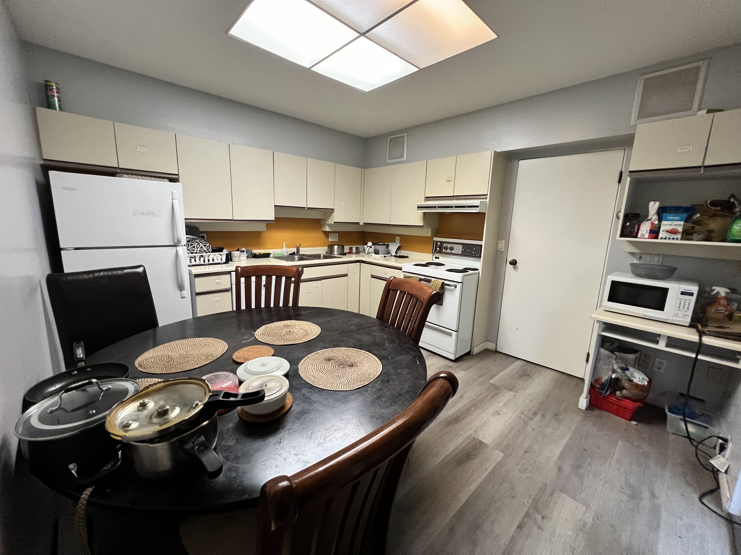 A kitchen with beige cabinets, a white refrigerator, a white stove, a microwave, and a black dining table with four wooden chairs. The table has several placemats and some kitchen items on it.