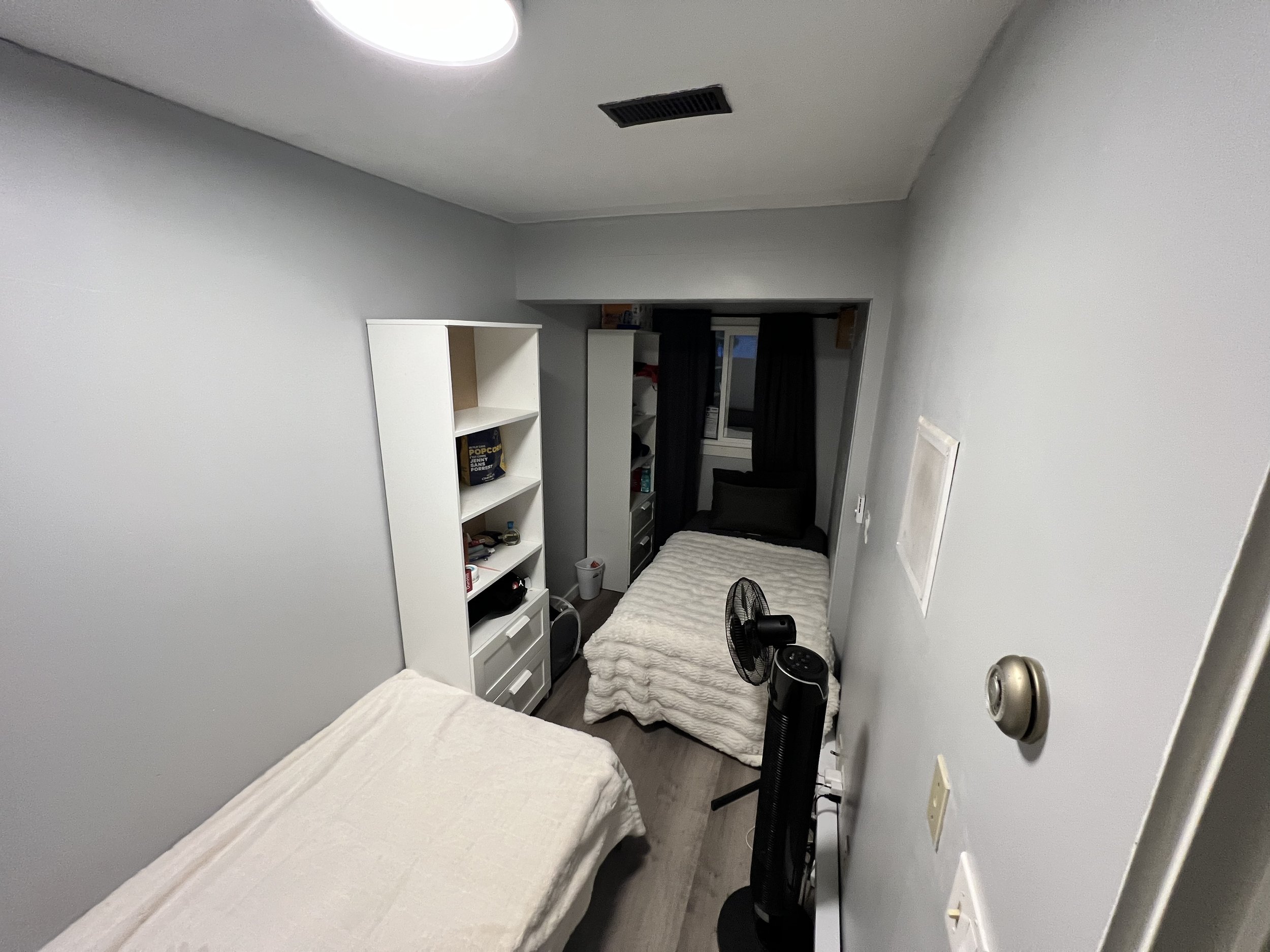 A small bedroom with two beds, white blankets, a black fan, a tall white bookshelf, and a window with black curtains.