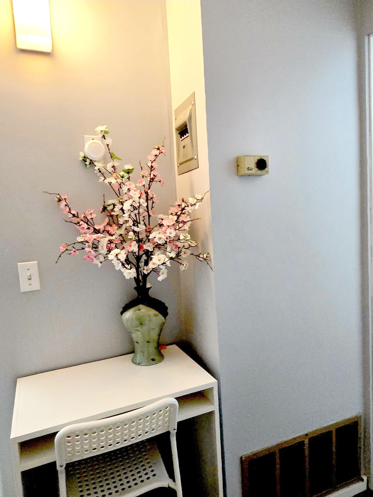 A corner of a room with a white desk, a white perforated chair, and a ceramic vase with pink and white cherry blossom branches placed on the desk. The room has light gray walls with a thermostat, a light switch, and an intercom panel.