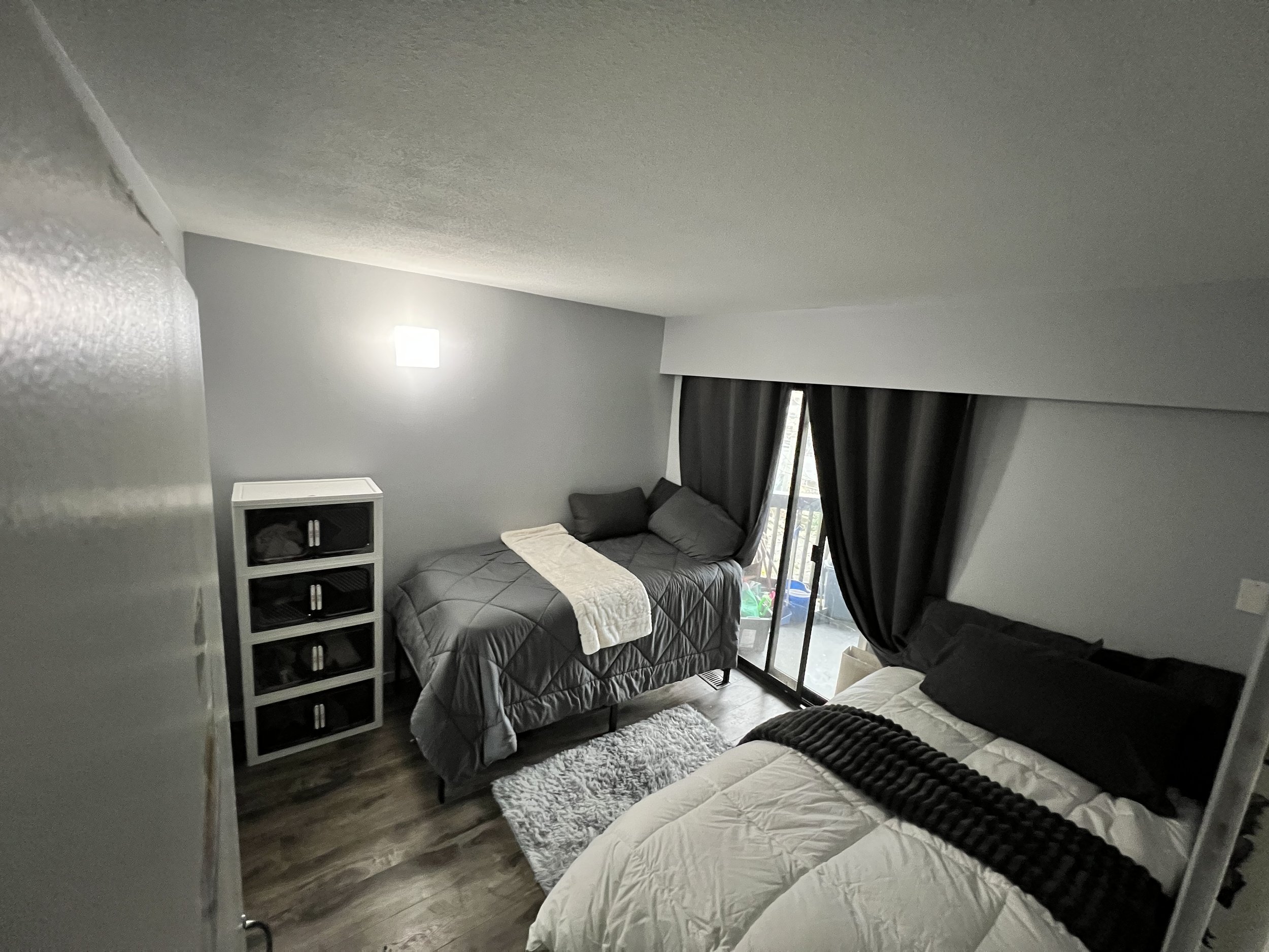 A small bedroom with two beds, a sliding glass door with black curtains, a white shelf, a wall-mounted light, and wooden flooring.