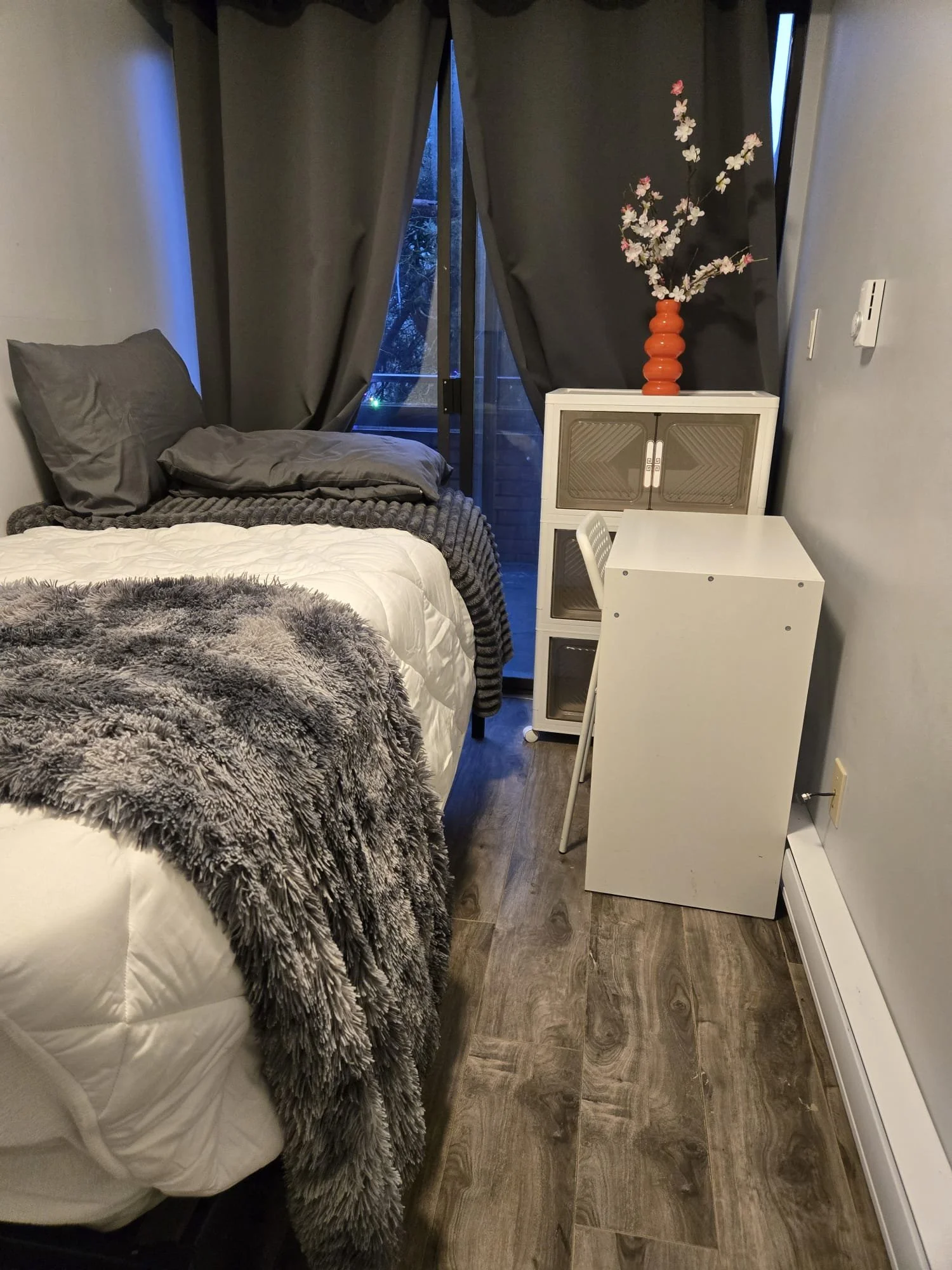 A bedroom with a bed dressed with dark gray and white bedding, a gray plush blanket, and matching pillows. Behind the bed, there's a sliding glass door with dark gray curtains, leading to an outdoor space. On the right side, a white cabinet with a decorative orange vase holding pink and white flowers on top. In front of the cabinet, a small white table with a white chair. The flooring is wood, and the walls are painted light gray.