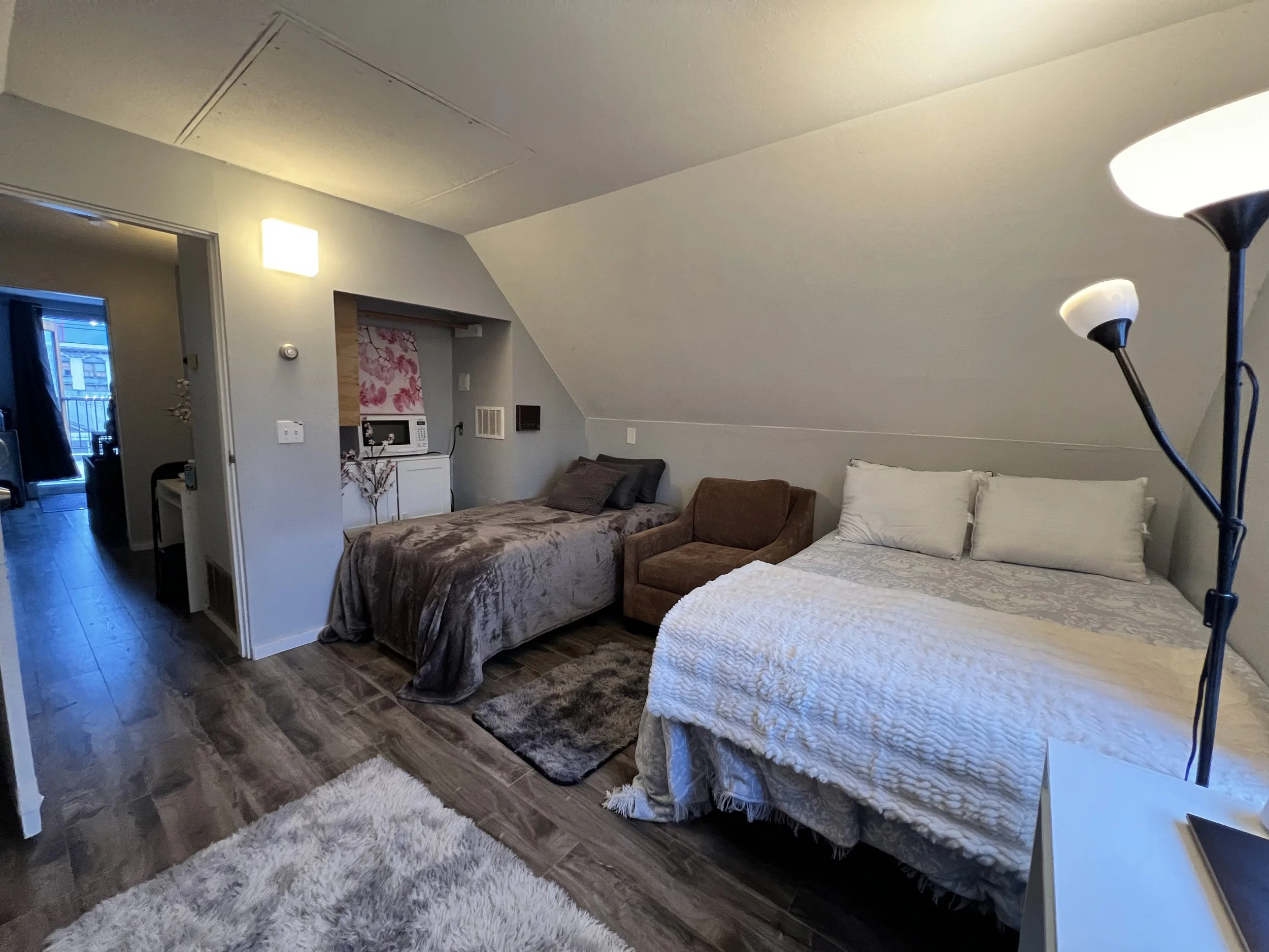 A cozy attic bedroom with sloped ceiling, featuring a bed with white bedding, a brown armchair, a brown bed with gray pillows, and a small white bedside table with a lamp. The room has hardwood flooring, a fluffy white rug, and a floor lamp.