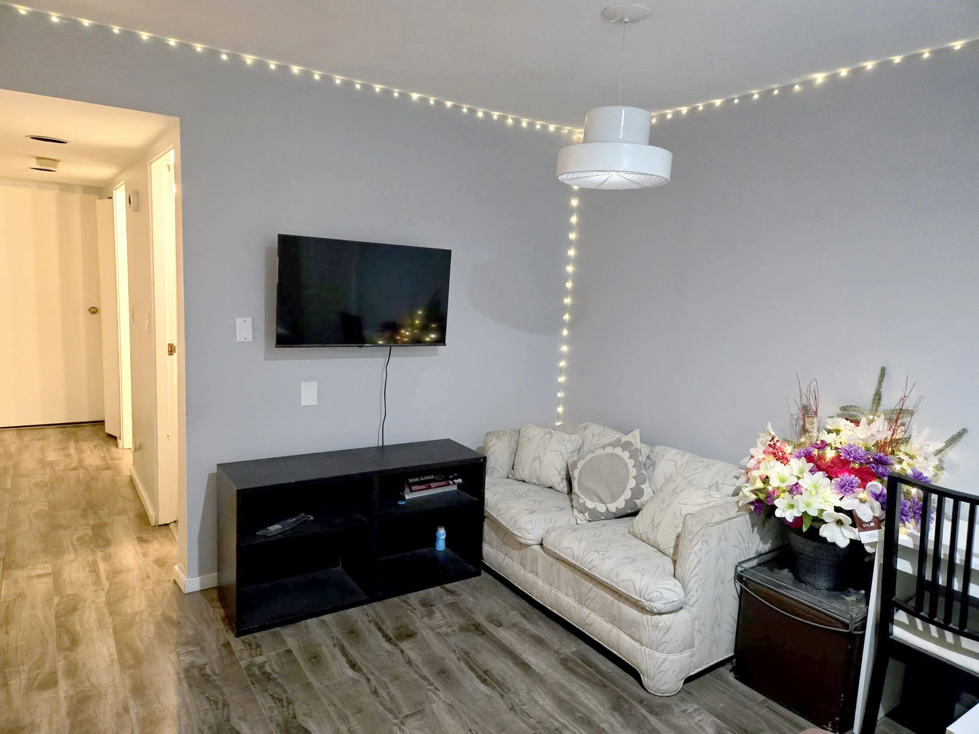Living room with a flat-screen TV mounted on light gray wall, black TV stand, cream sofa with decorative pillows, large floral arrangement on a small table, string lights along the ceiling and wall, and a modern ceiling lamp.