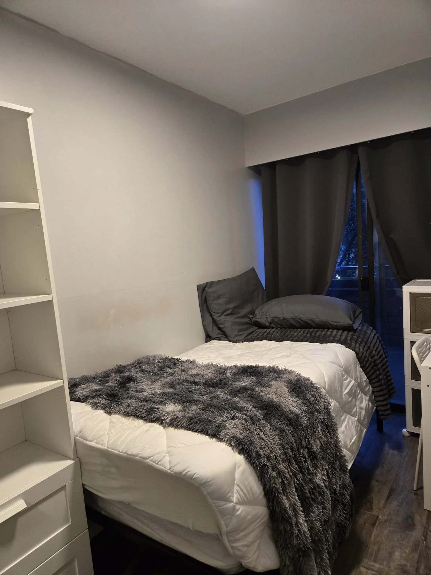A cozy bedroom with a bed covered in white quilt, gray and black pillows, and a plush gray faux fur throw blanket. There's a white bookshelf on the left and dark curtains partially covering a sliding glass door leading outside. The floor appears to b