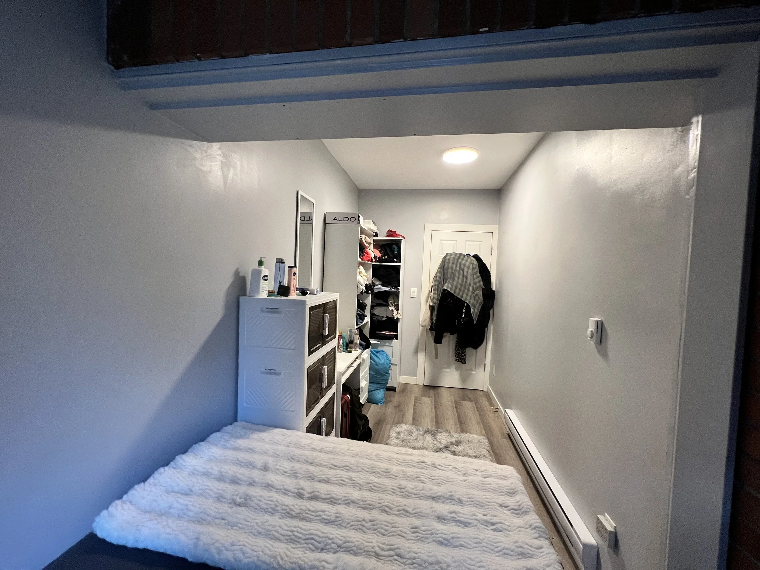 A small bedroom with white walls, a bed with a white textured blanket, a white storage cabinet with bottles and personal items on top, a tall open shelf with clothing, and a white door with clothes hanging on it. The room has a wooden floor and a cei