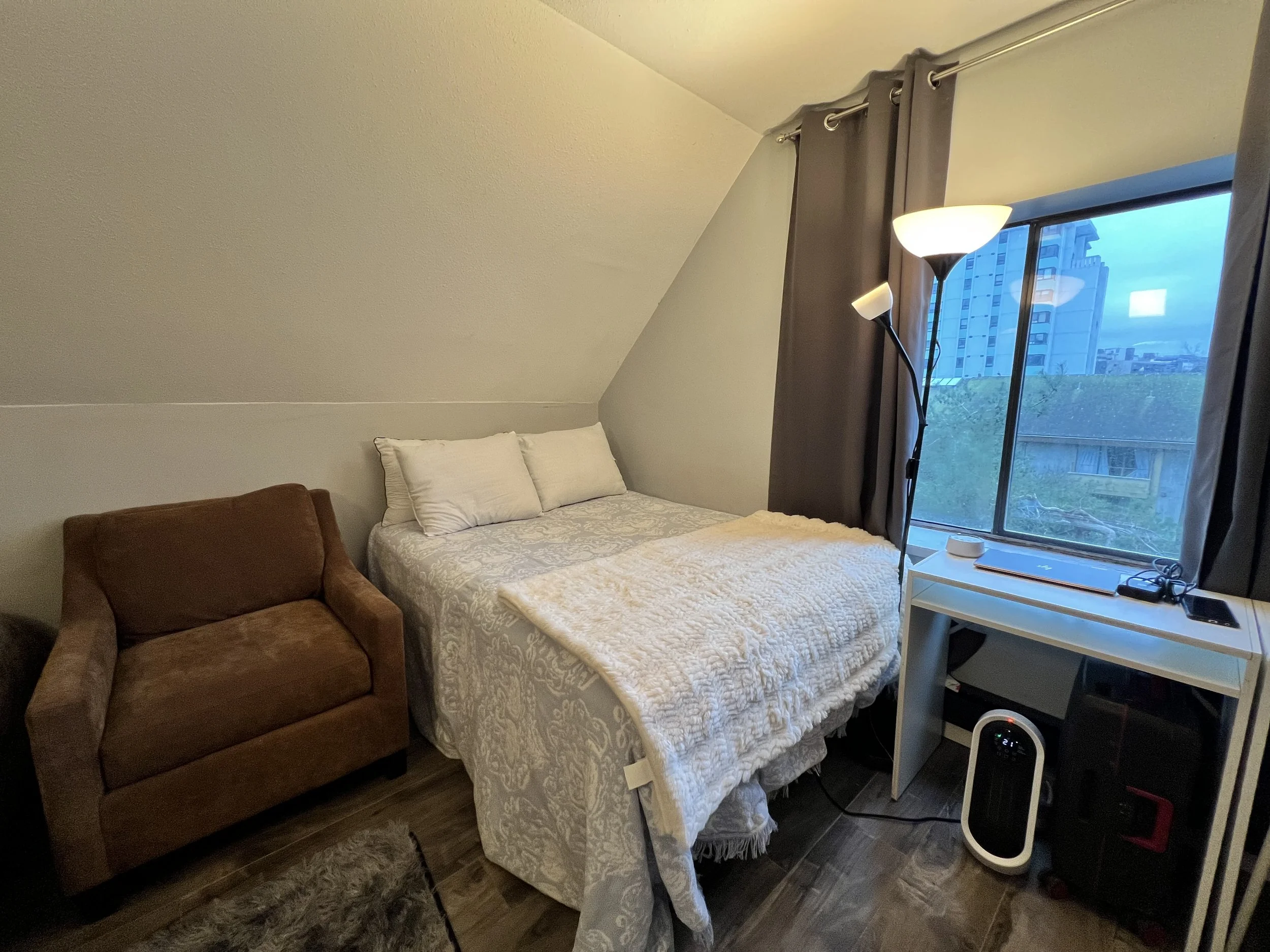 Small bedroom with a slanted ceiling, featuring a bed with white bedding and pillows, a brown armchair, a white side table with a lamp and some items, a window with dark curtains, and a floor fan near the window, with a cityscape visible outside.