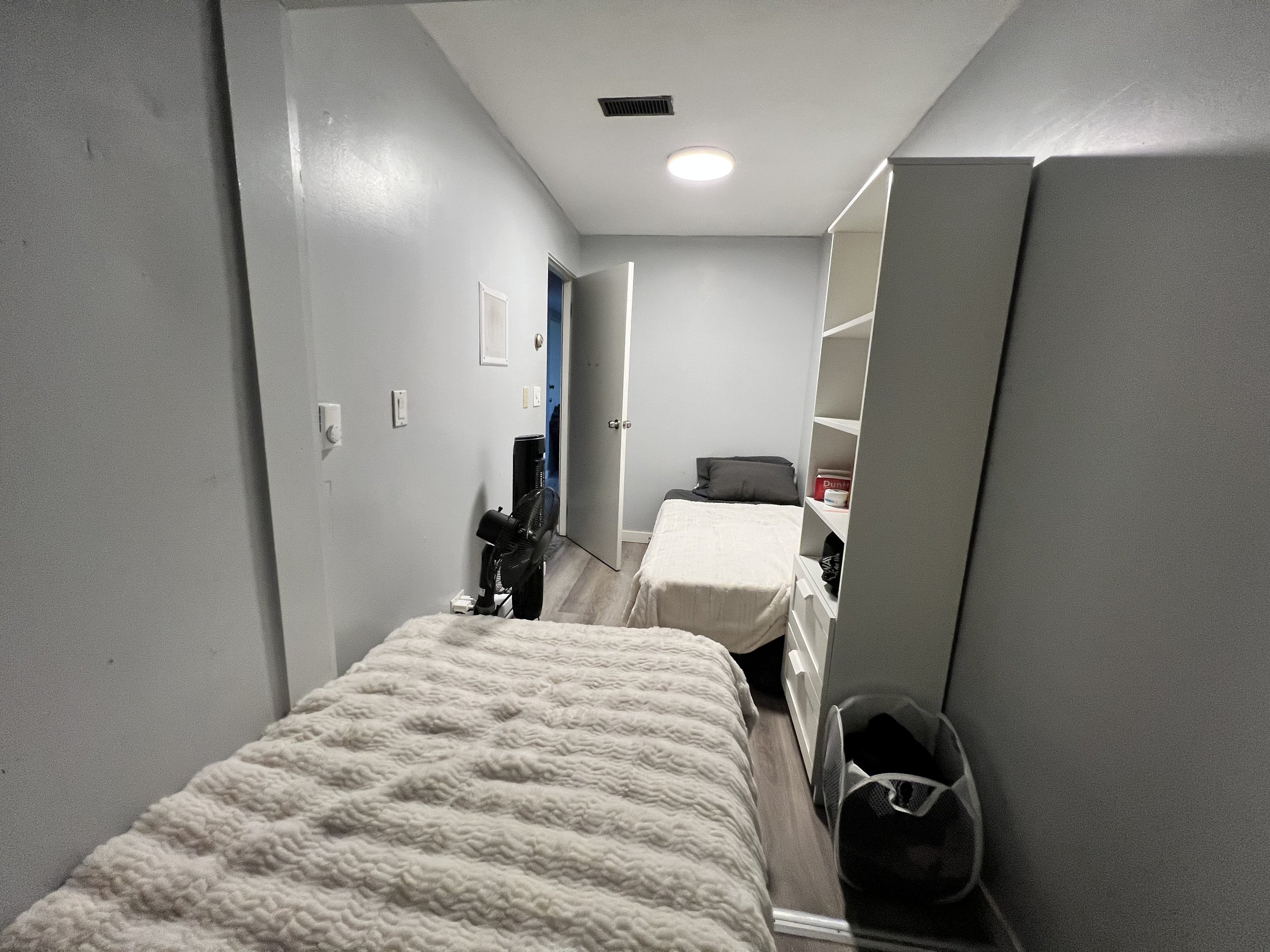 A small, simply furnished bedroom with two beds, a bookshelf, and a laundry basket, illuminated by a ceiling light.