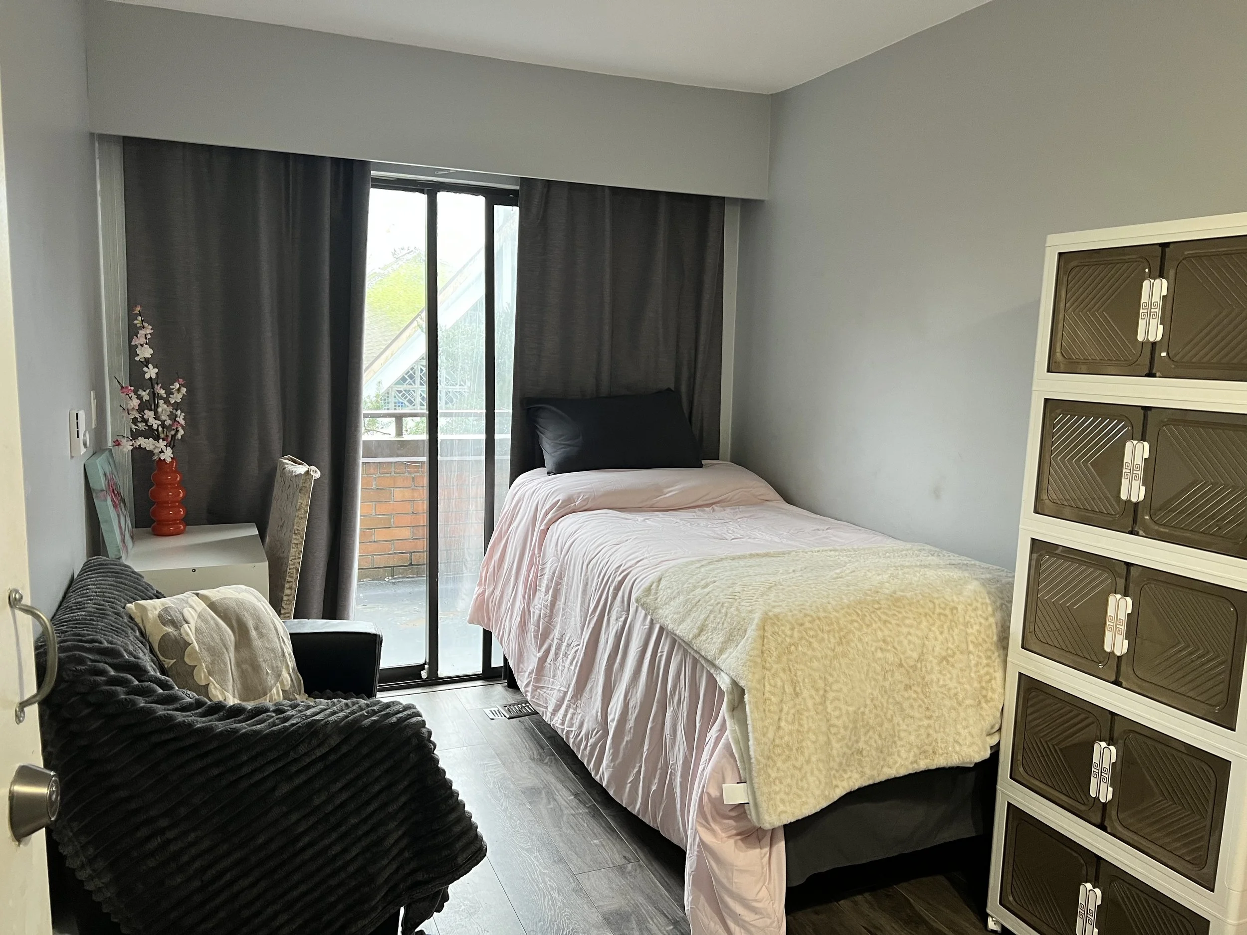 A bedroom with a bed near the sliding glass door, covered with a pale pink blanket and a beige throw blanket, black pillow, dark curtains, a black sofa with beige cushion, a small white desk with a red vase and artificial flowers, and a brown and white plastic storage cabinet.