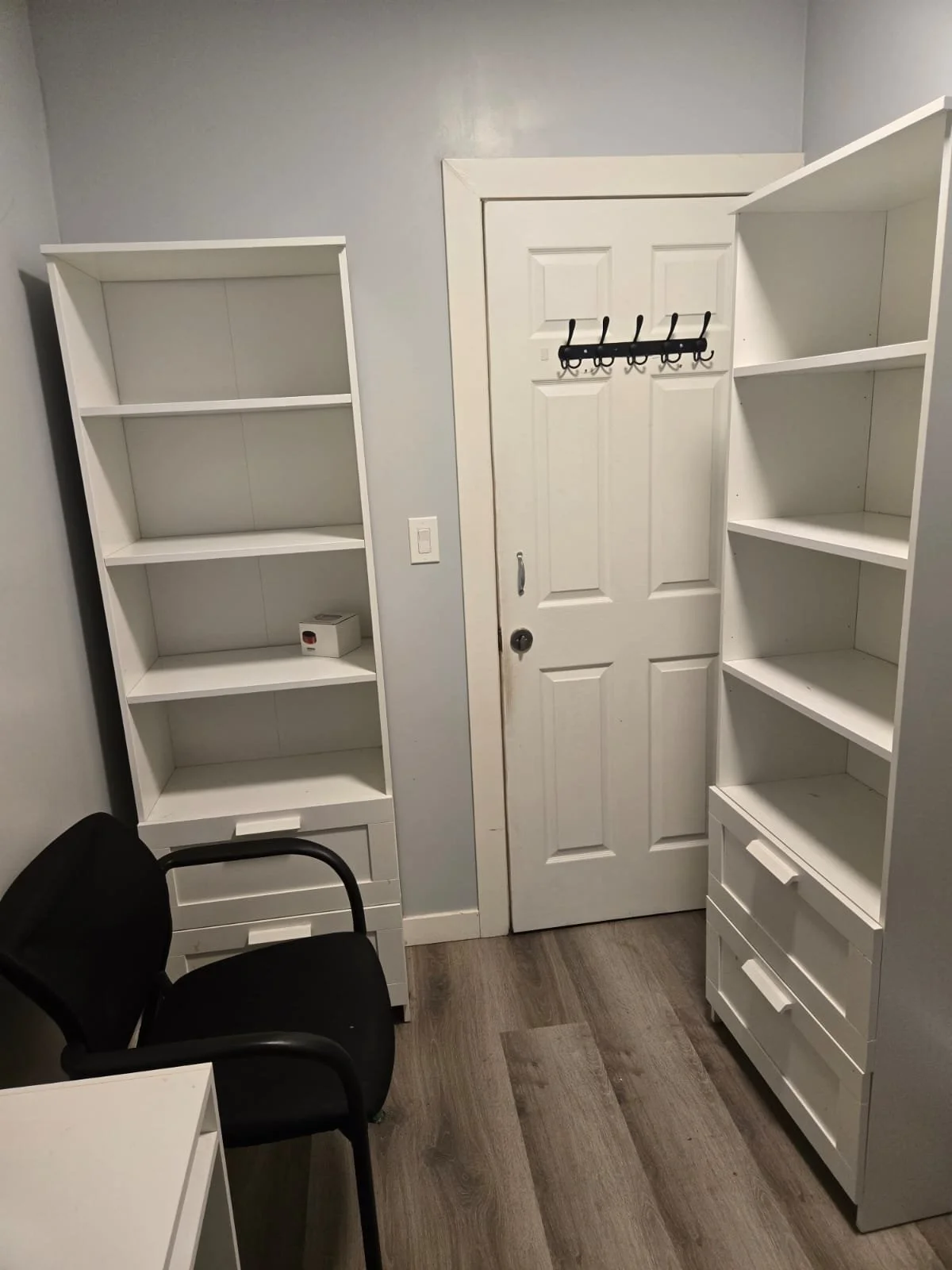 A small room with a white door, two white bookshelves, a black chair, and a white desk.