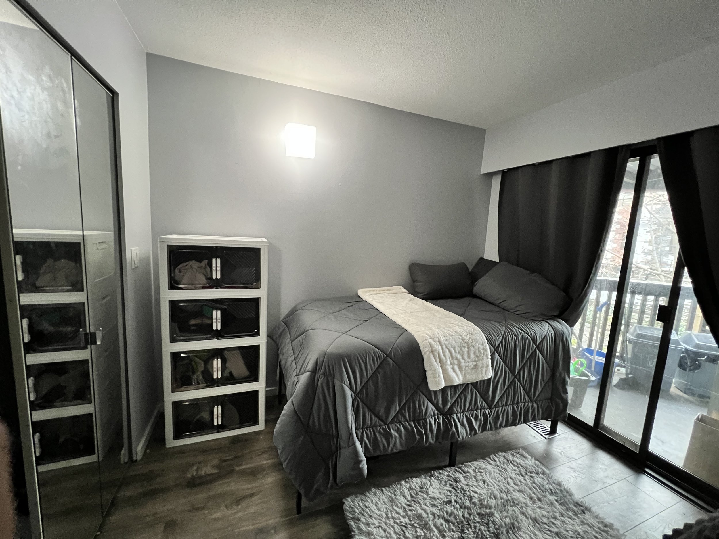 Small bedroom with gray bedding, black curtains, sliding glass door leading to a balcony, and a white storage unit.