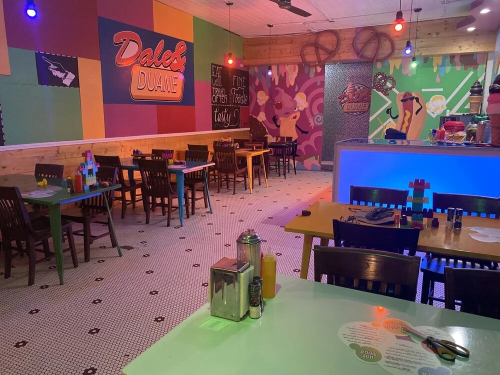 Colorful interior of a casual restaurant or diner with comic-style wall art, wooden accents, and small tables set for dining, featuring LEGO creations as table decorations.