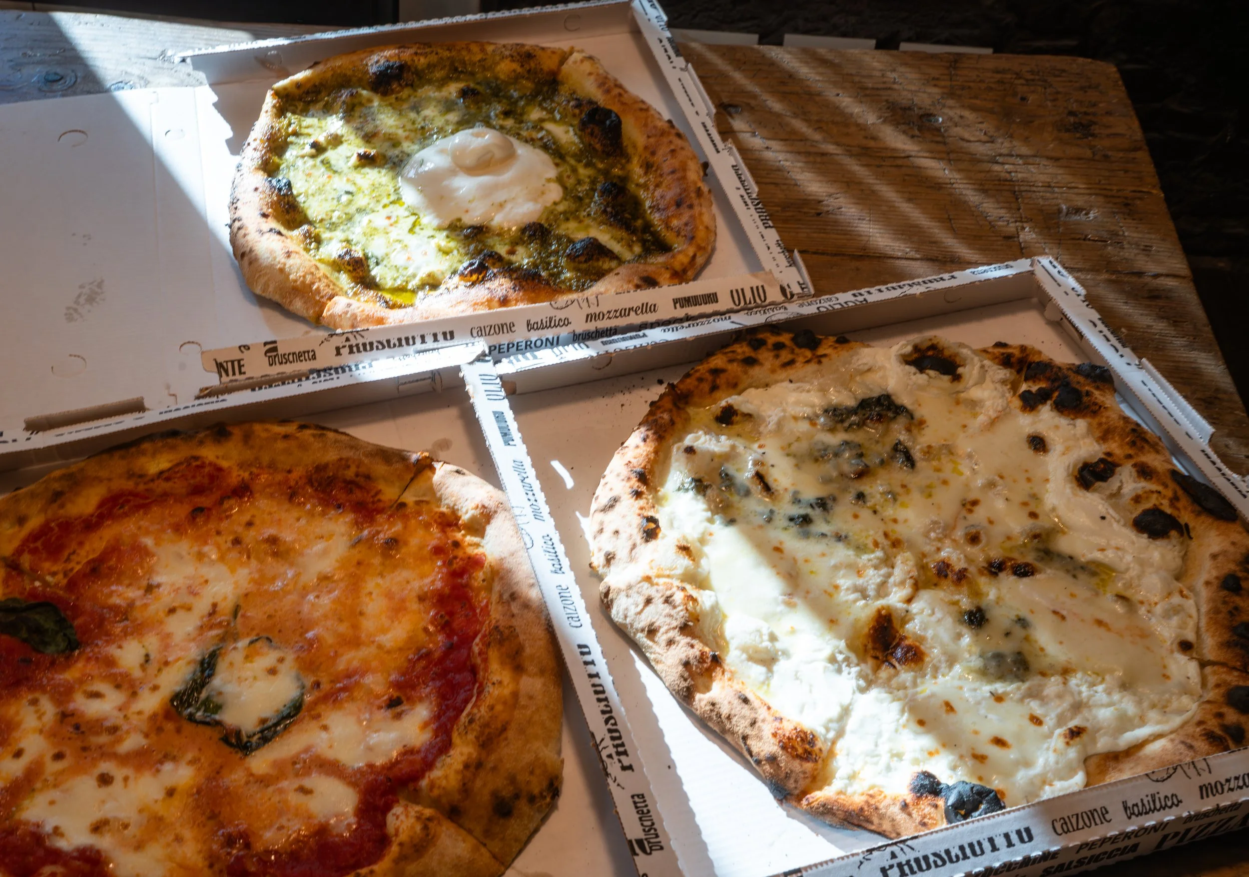Three different pizzas in open pizza boxes on a wooden table, with the top pizza topped with an egg, the bottom right pizza with cheese and black truffle, and the bottom left pizza with cheese, tomato sauce, and basil.