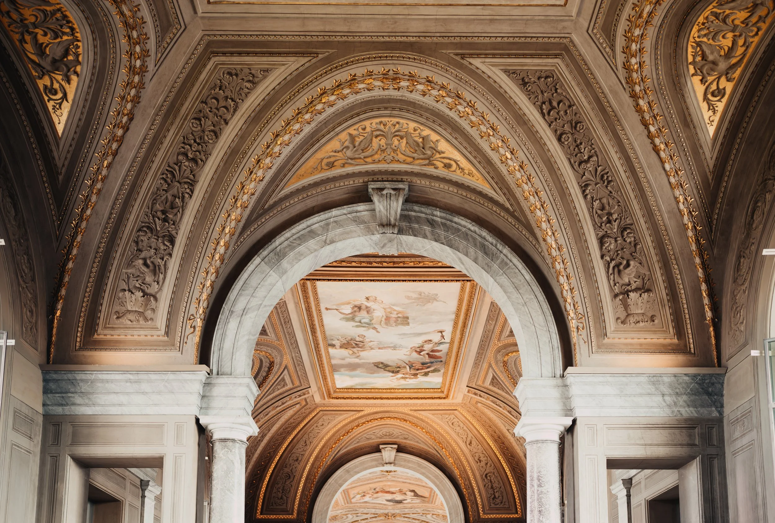 Interior of a grand building with ornate arches, intricate gold and white ceiling details, and classical painting on ceiling.