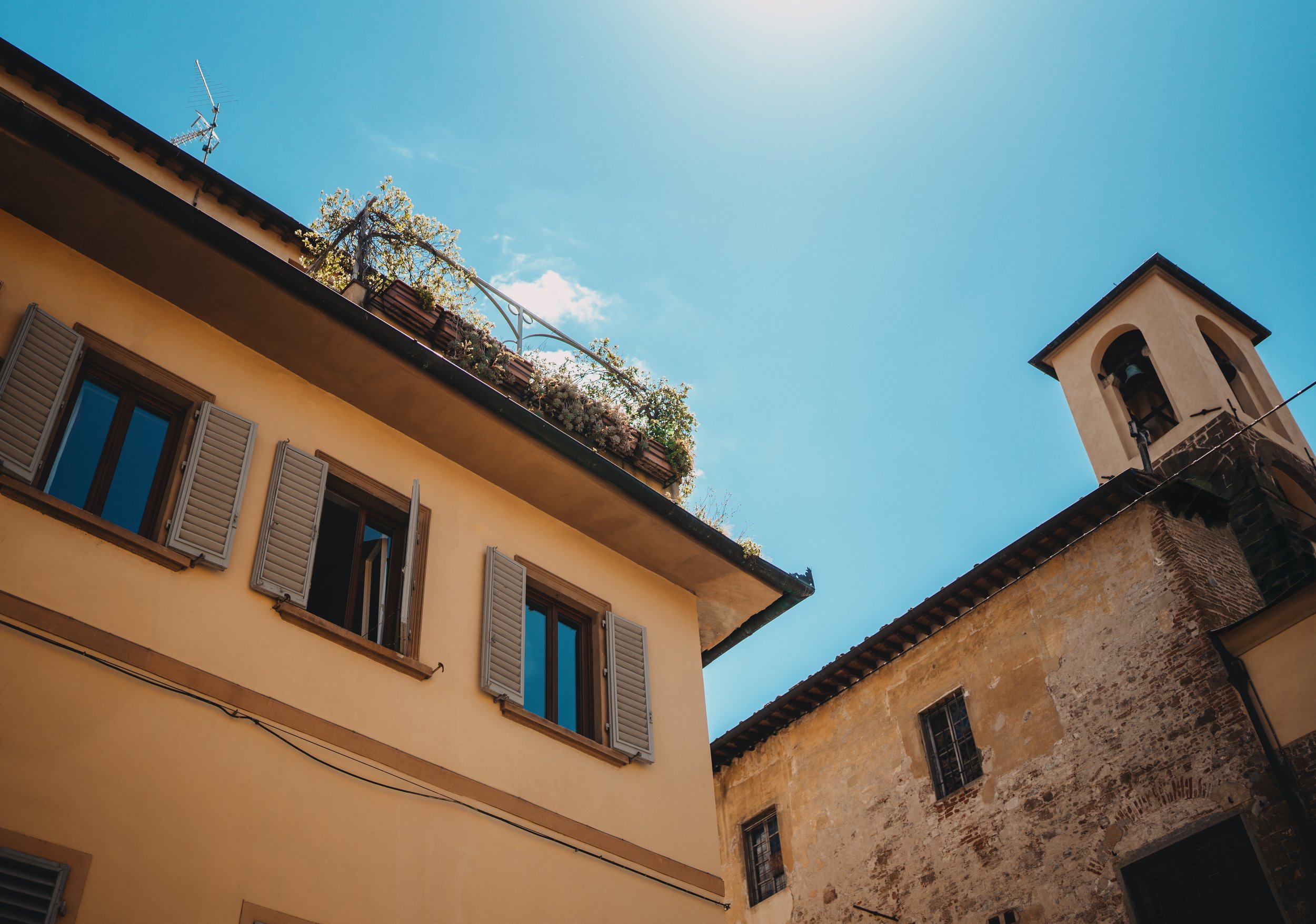 A view looking up at old European buildings with shutters and a church bell tower against a bright blue sky.
