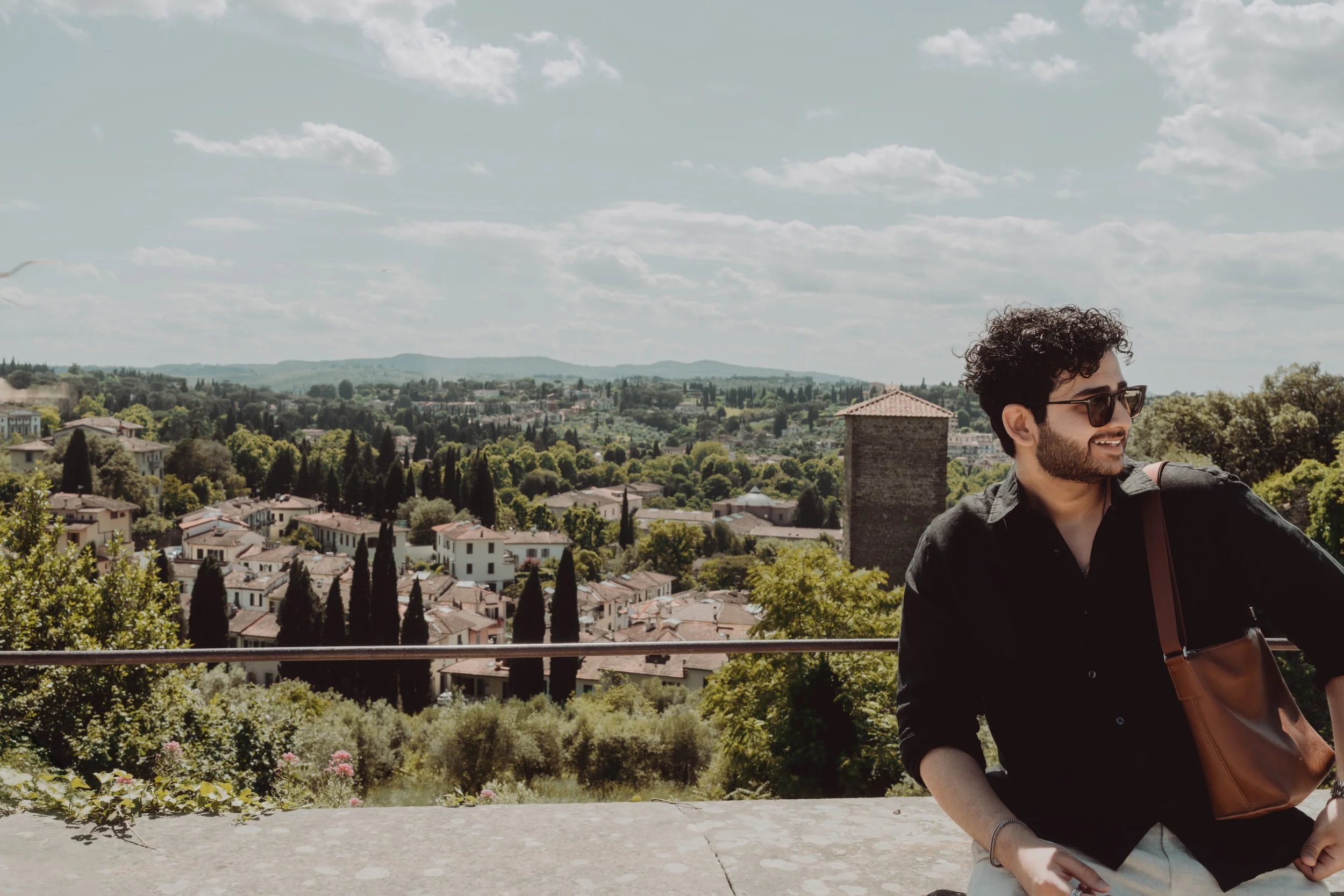 Young man with curly hair, wearing sunglasses and a black shirt, sitting outdoors with a cityscape and green trees in the background.