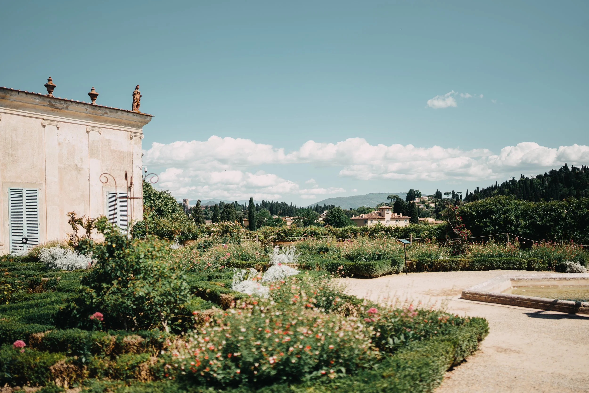 A scenic landscape with a garden of rose bushes and a rustic building on the left, under a sky with scattered clouds and distant hills in the background.