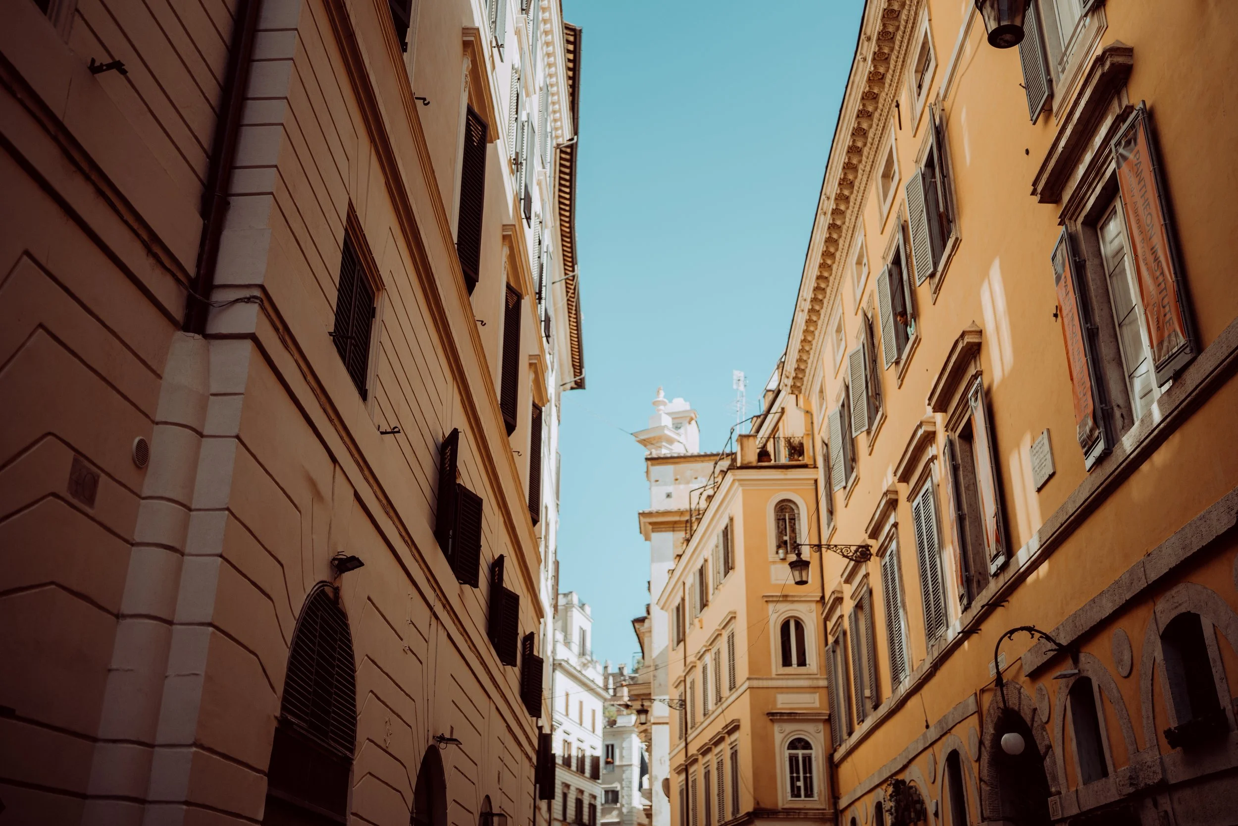 Tall narrow buildings with shuttered windows and a cafe sign along a European street, with blue sky overhead.