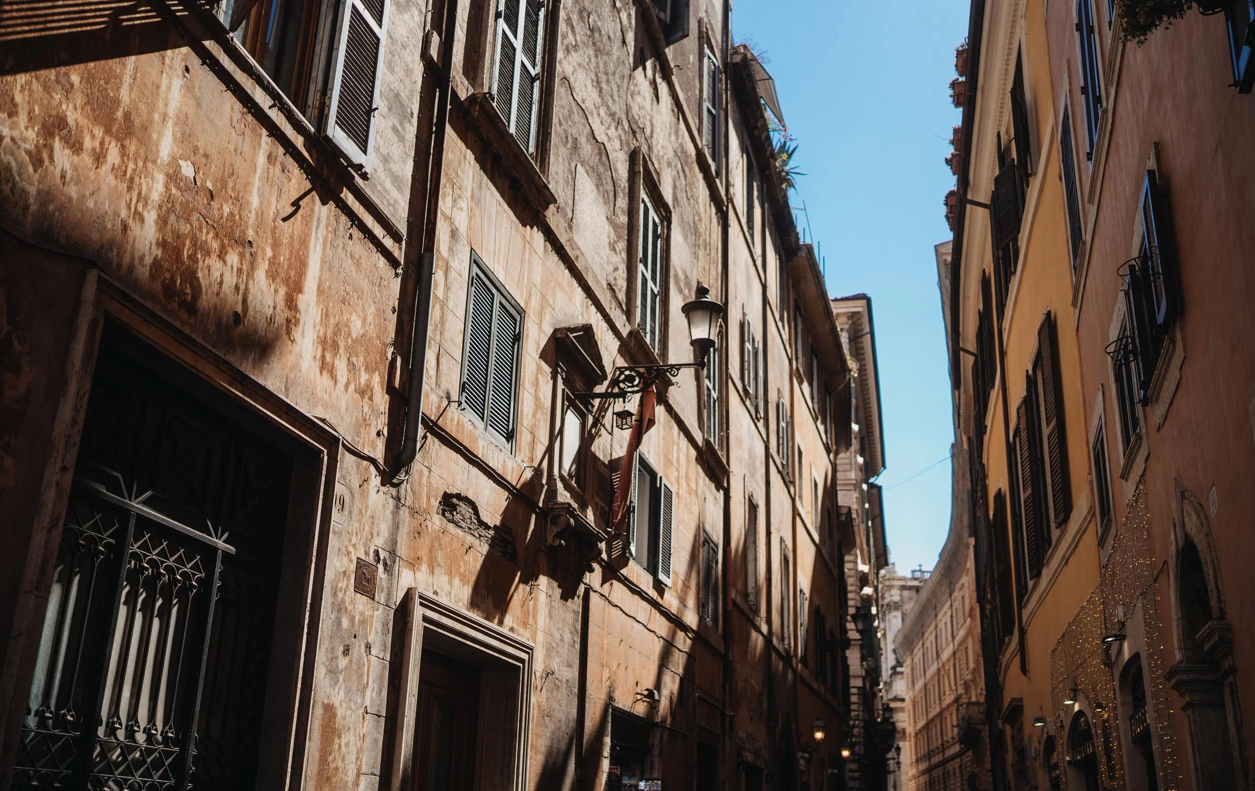 Narrow European street with historic buildings, some with peeling paint, black shutters, and a streetlamp, under a clear blue sky.