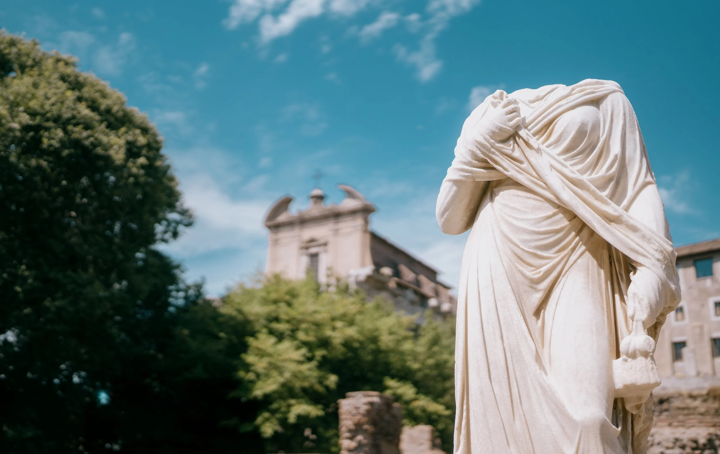 A white marble statue of a headless woman with draped clothing holding a handkerchief, set against a background of green trees and historic stone buildings under a blue sky.
